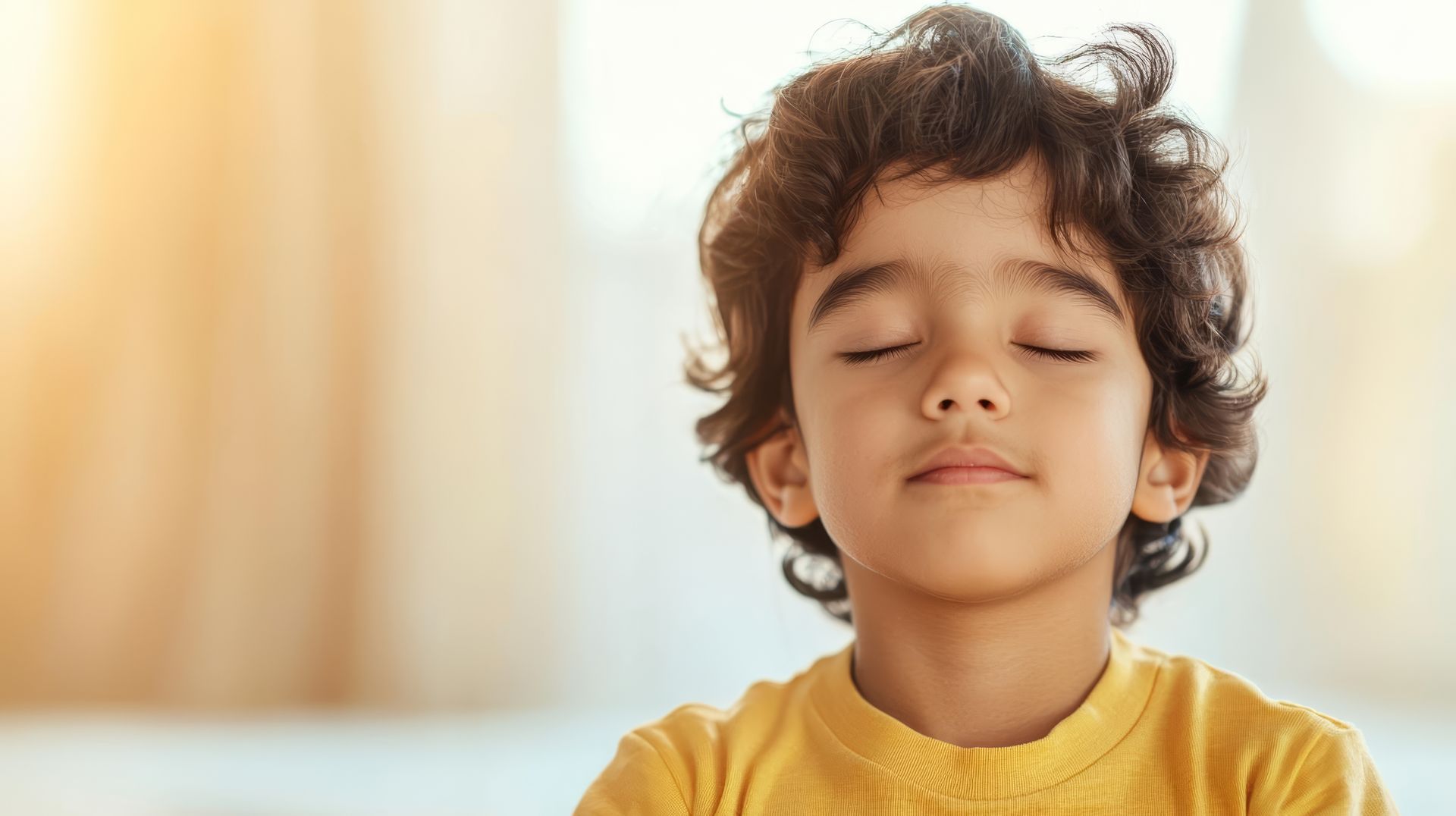 A child with curly hair wearing a yellow shirt, sitting with eyes closed in a peaceful, bright indoor setting.