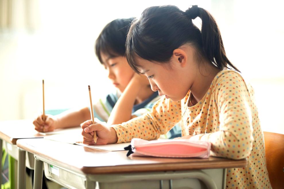 Two students sit at desks in a bright classroom, intently focused on writing in their notebooks with pencils.