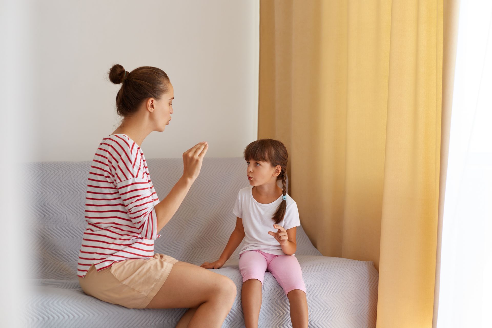 A woman in a striped shirt sits on a sofa, gesturing while speaking to a girl in a white shirt and pink leggings.