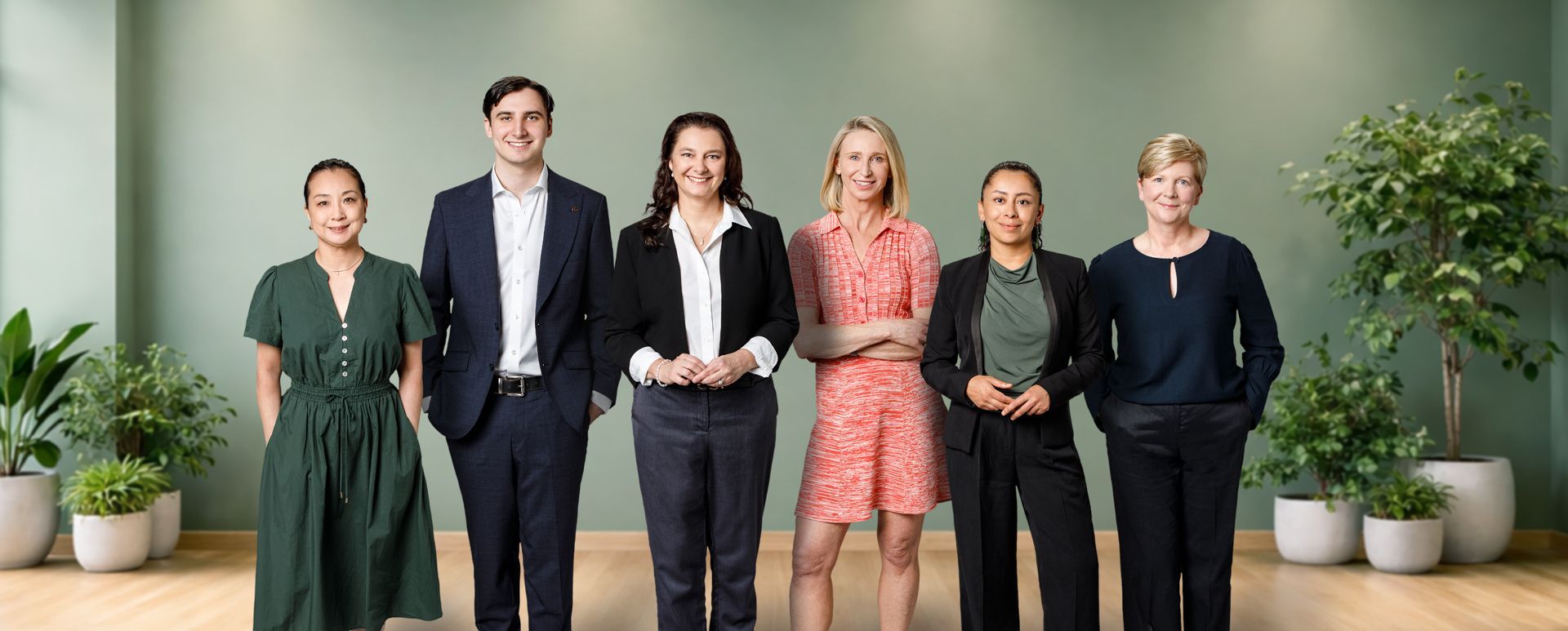 Six people stand in a row against a solid green wall in a bright office space with potted plants, smiling at the camera.