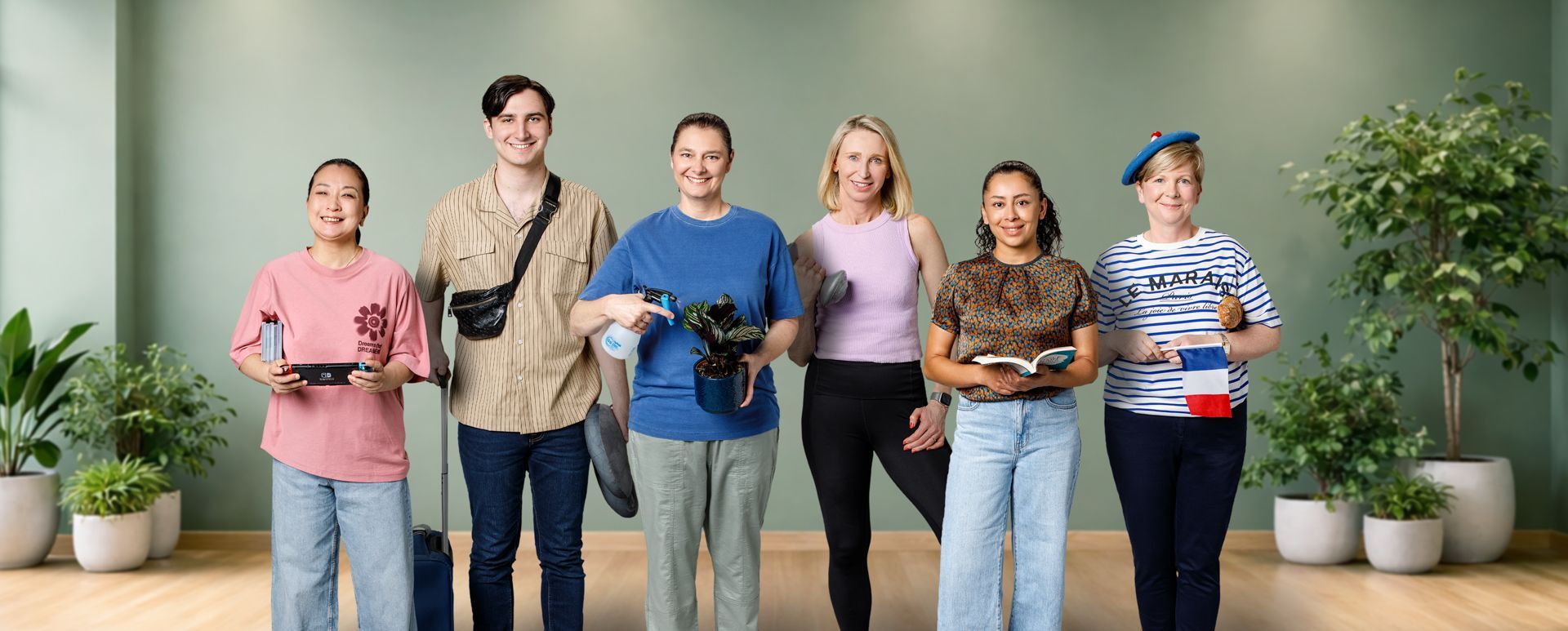 Six people stand in a row against a light green wall with potted plants, smiling and holding travel-related items.