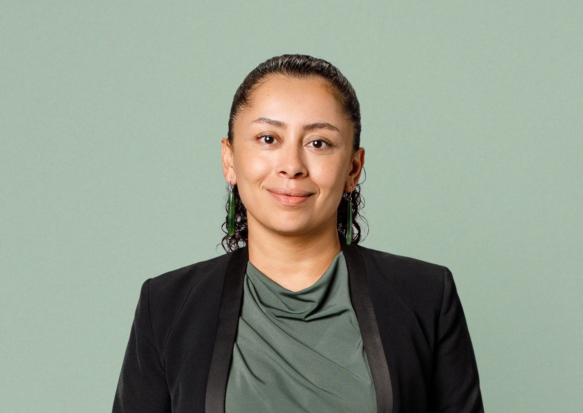A person with dark curly hair wearing a dark blazer over a green top, smiling against a light green background.