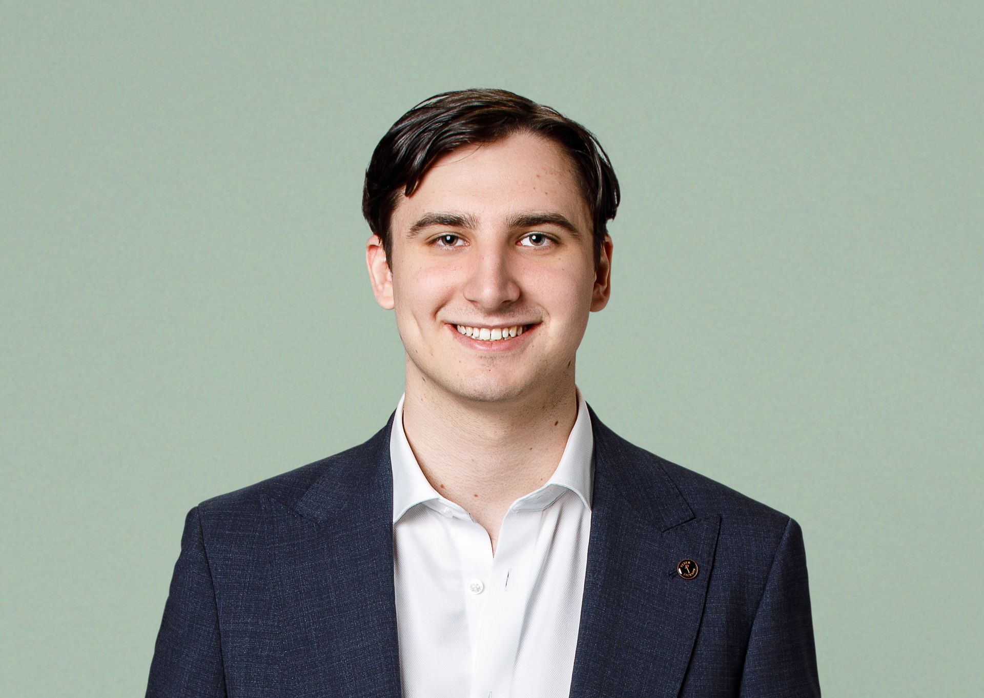 A professional headshot of a smiling person wearing a white collared shirt and a dark navy blazer against a sage green wall.