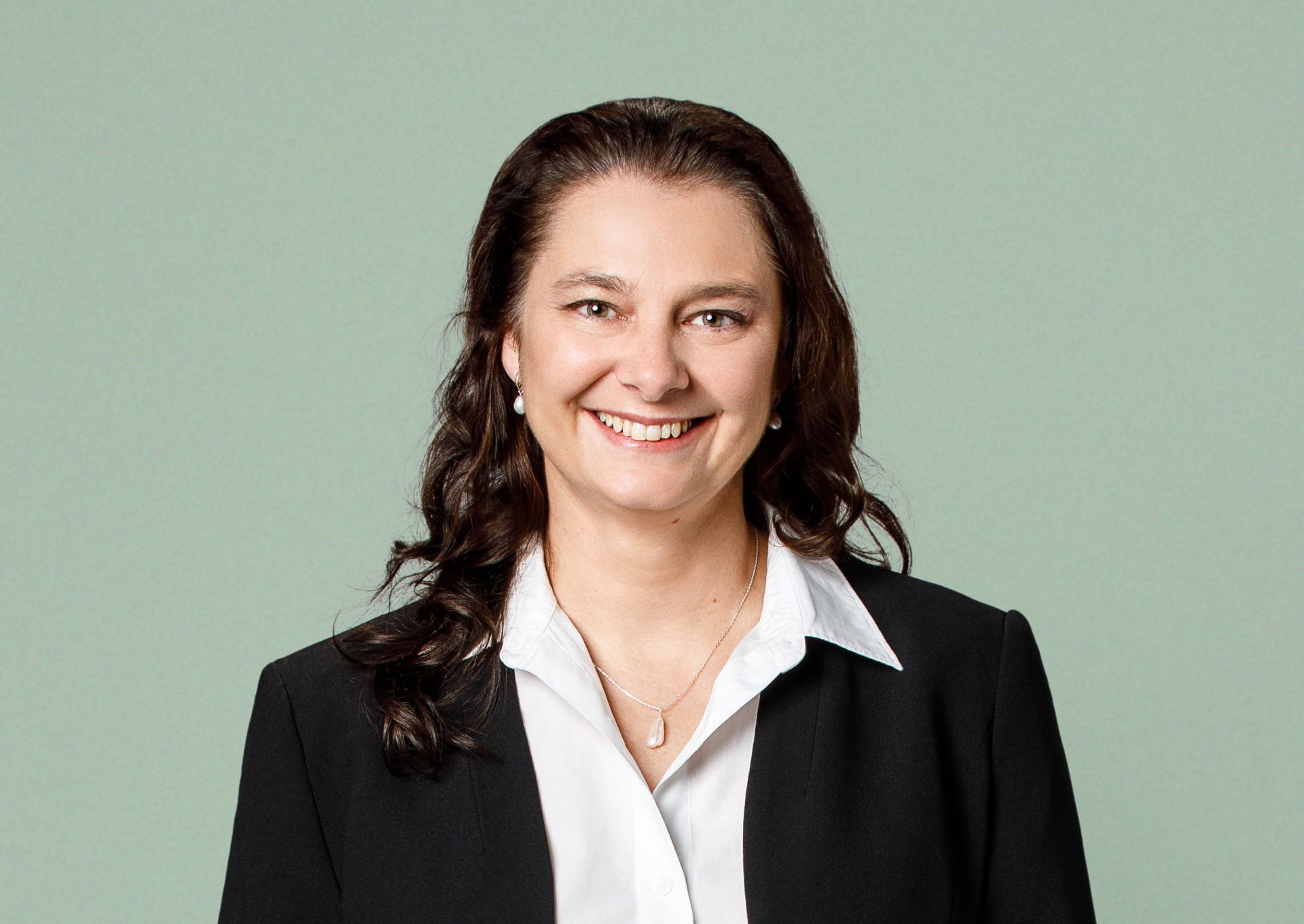 A smiling person with long brown hair wearing a dark blazer and white collared shirt against a light green background.