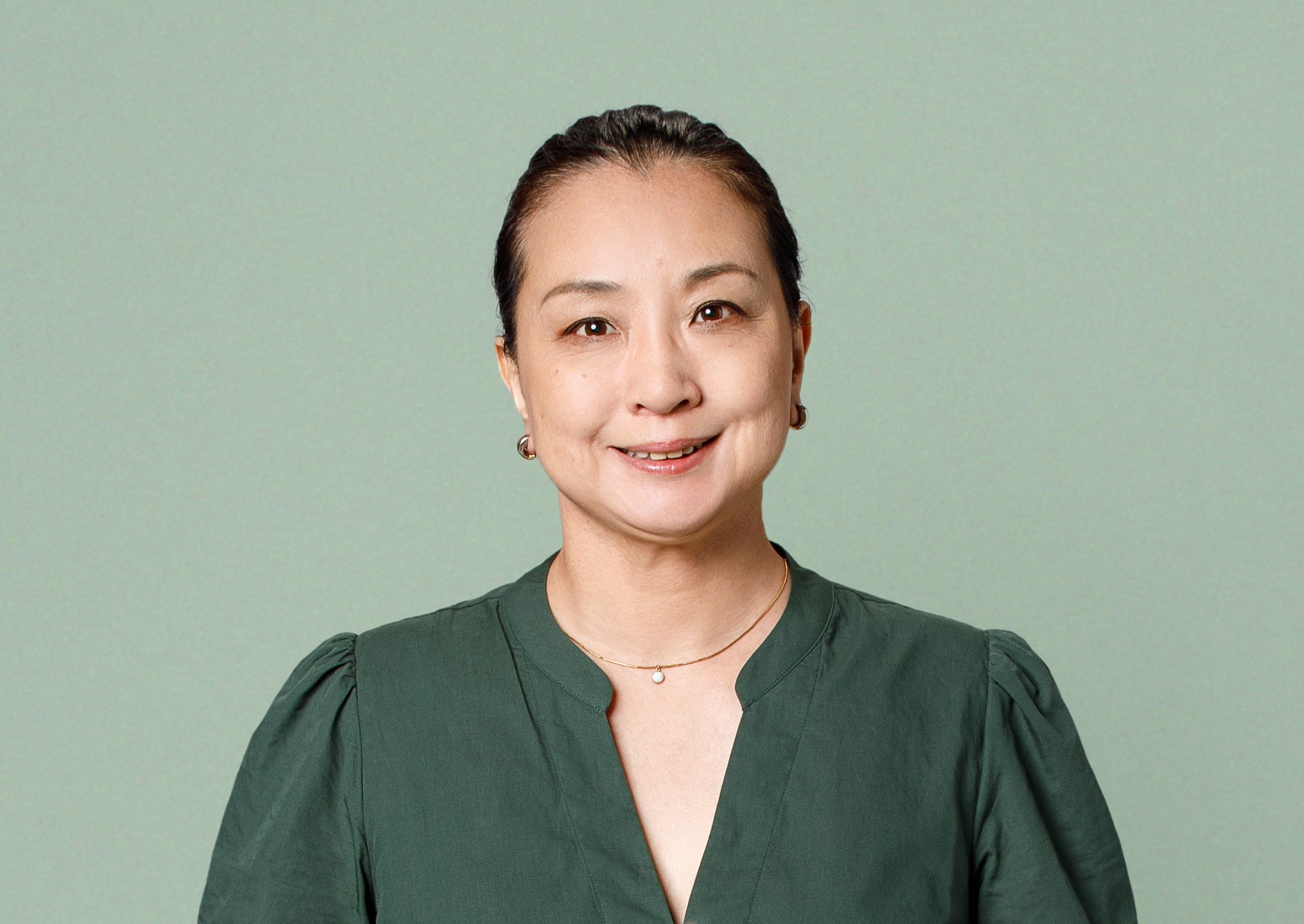 Smiling person with dark hair pulled back, wearing a dark green v-neck top and a small pendant necklace against a light background.