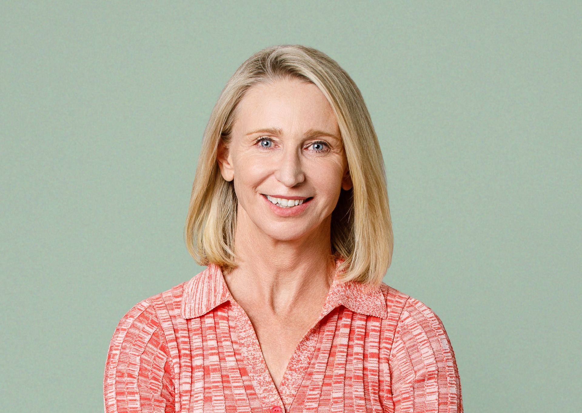 A person with blonde hair smiling, wearing a coral, textured button-down shirt against a pale green background.