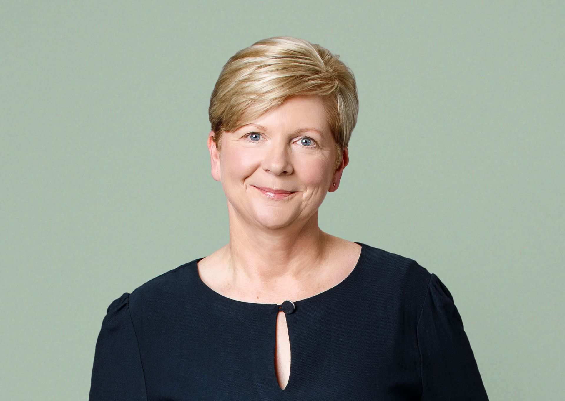 A person with short, light-brown hair and a gentle smile wearing a dark blue blouse against a pale green background.