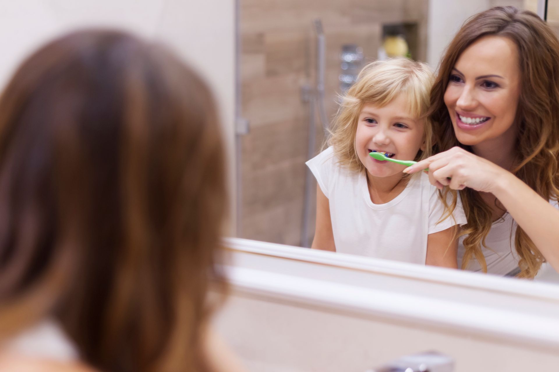 A person stands behind a child at a bathroom mirror, smiling while helping them brush their teeth with a green toothbrush.