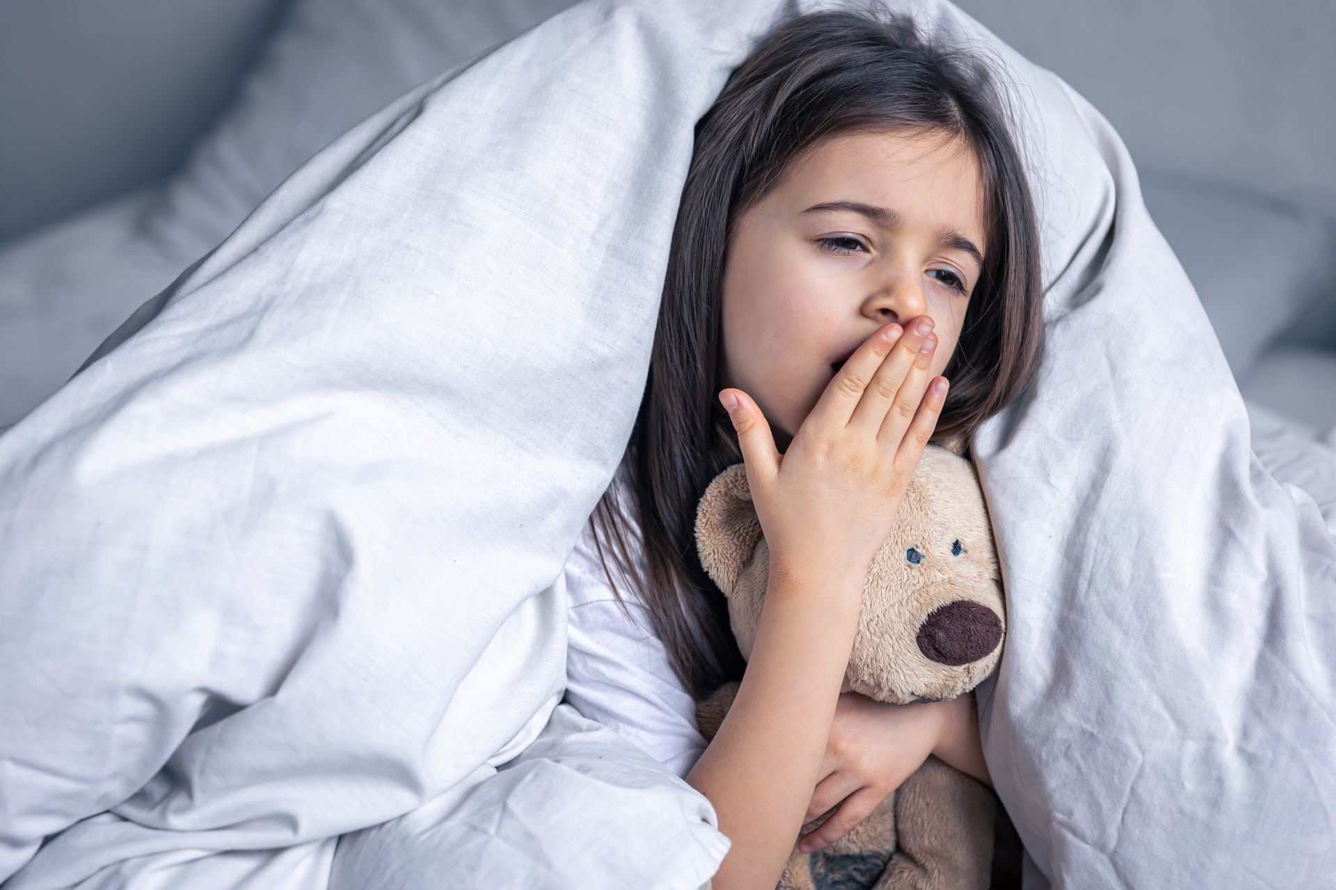 A child yawns while holding a teddy bear, wrapped in a soft white blanket in bed.
