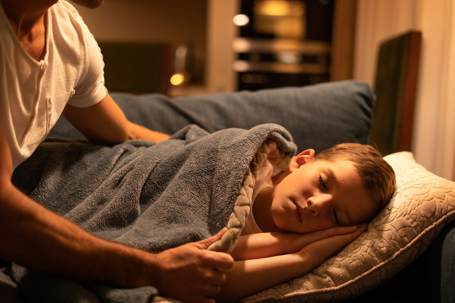 A person gently pulls a gray blanket over a child sleeping on a couch in a dimly lit room.
