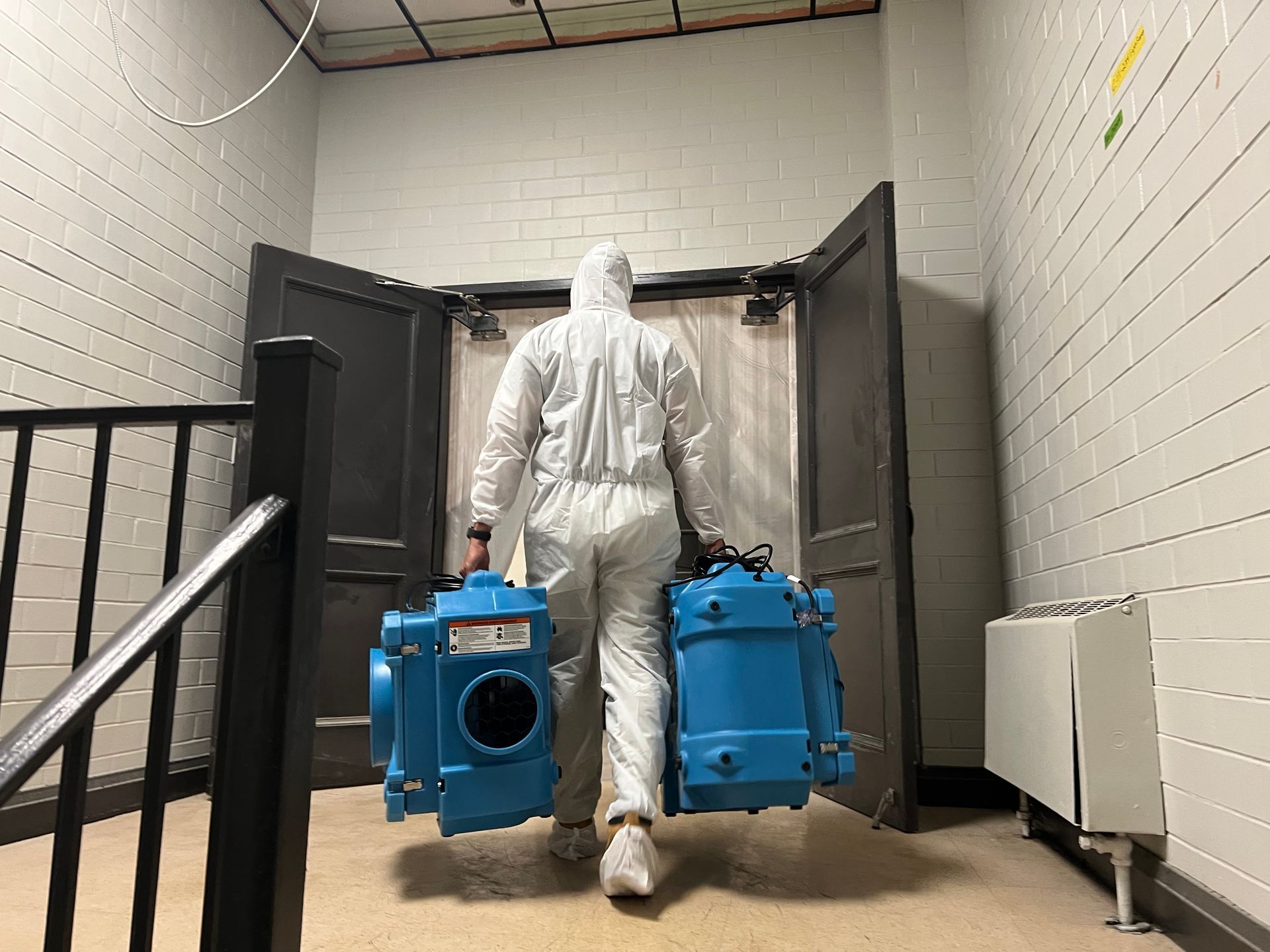 A man in a protective suit is carrying two blue boxes in a hallway.