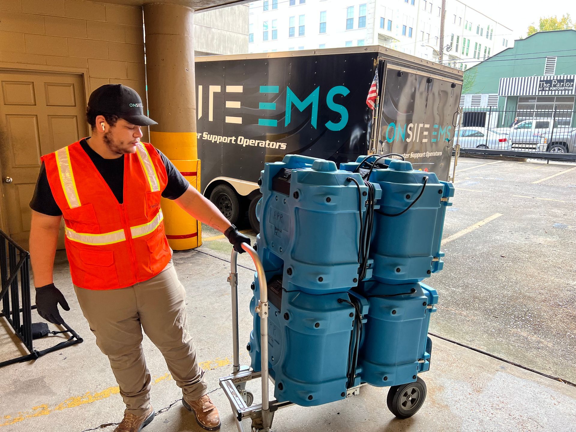 A man in an orange vest is pushing a cart full of blue barrels.