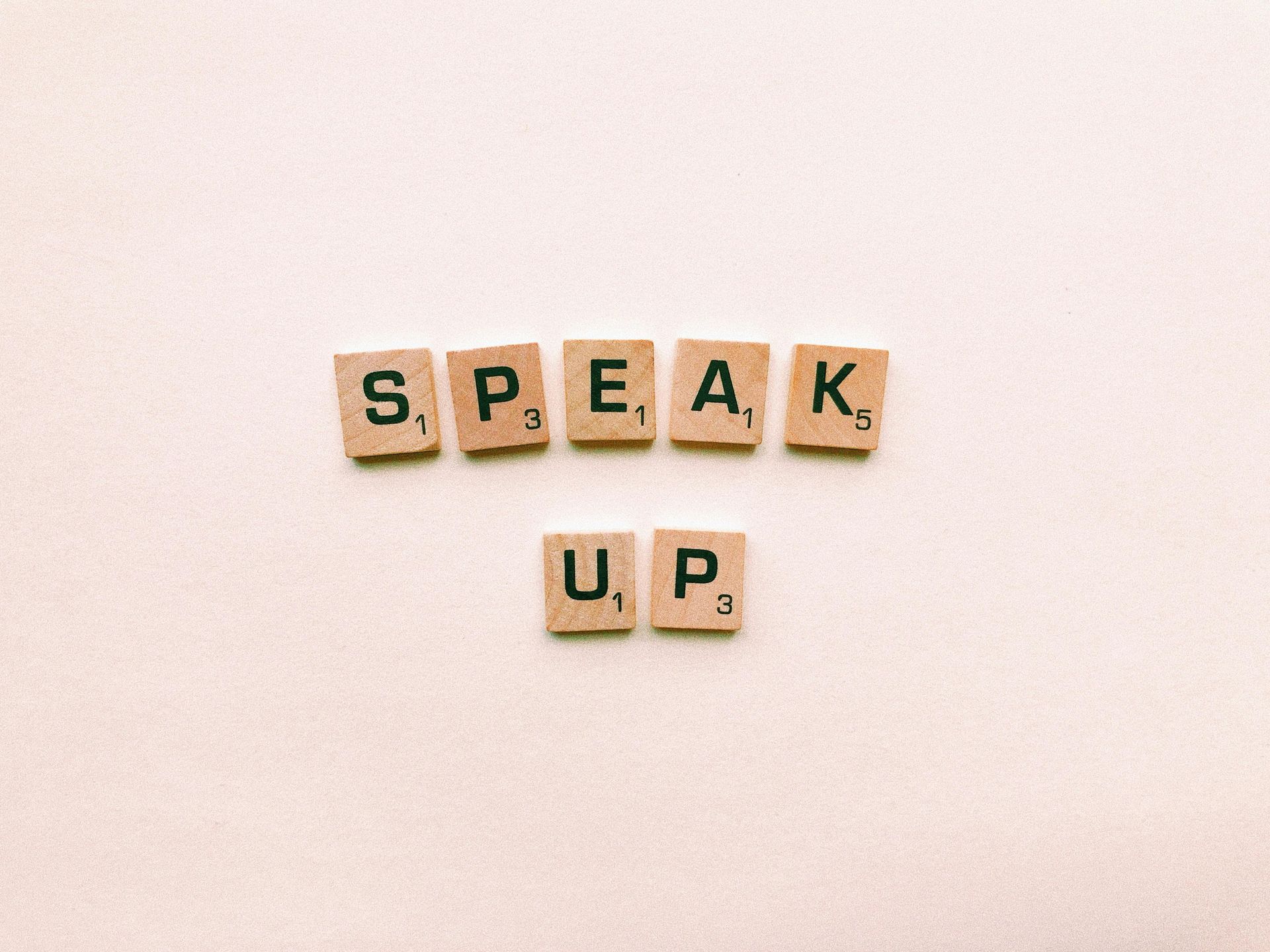 Scrabble tiles spelling the words “SPEAK UP” on a light beige surface, symbolizing communication and confident expression.