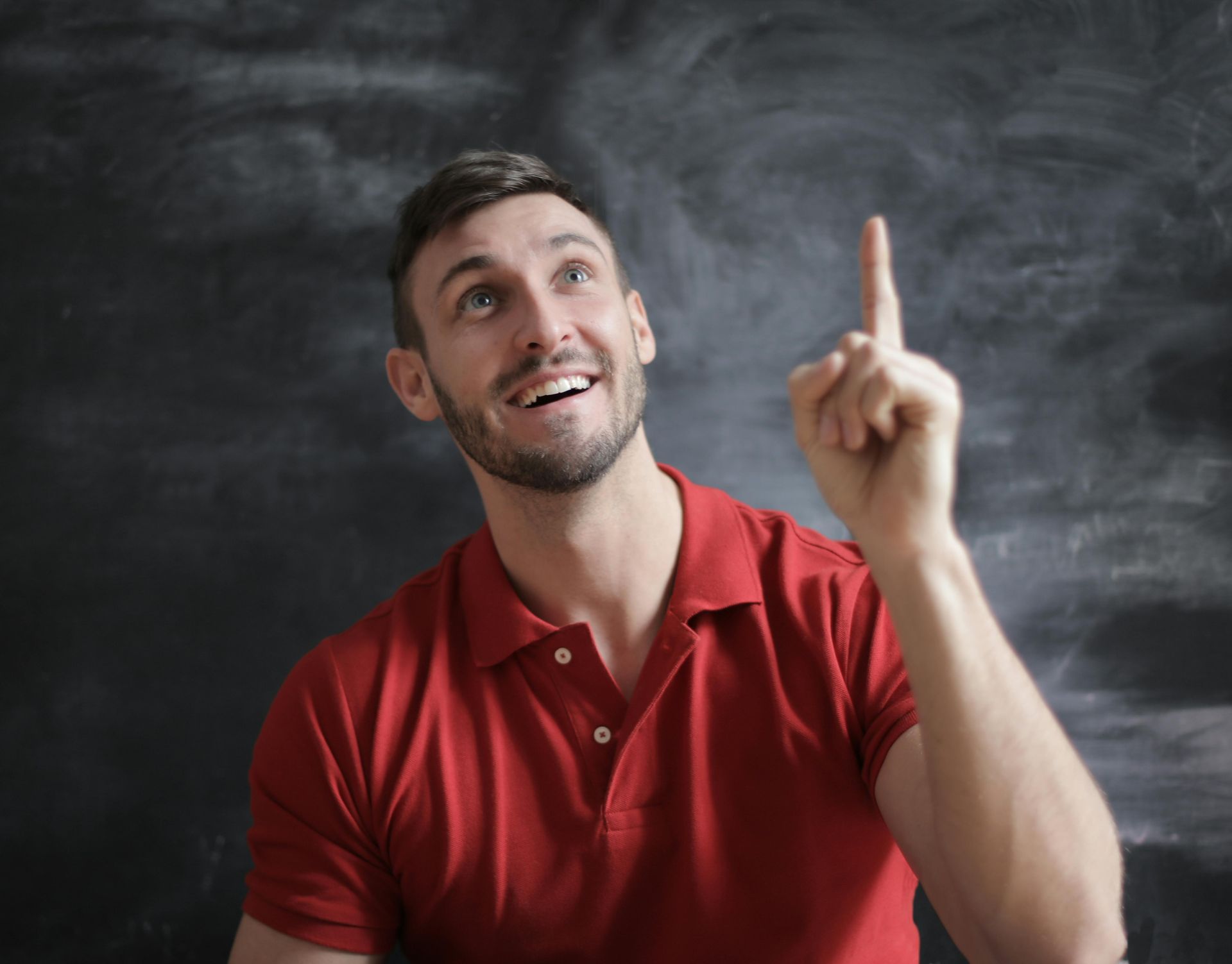 Smiling man in a red shirt pointing upward while sitting in front of a chalkboard, symbolizing inspiration, awareness, and the idea “What you think, you teach.”