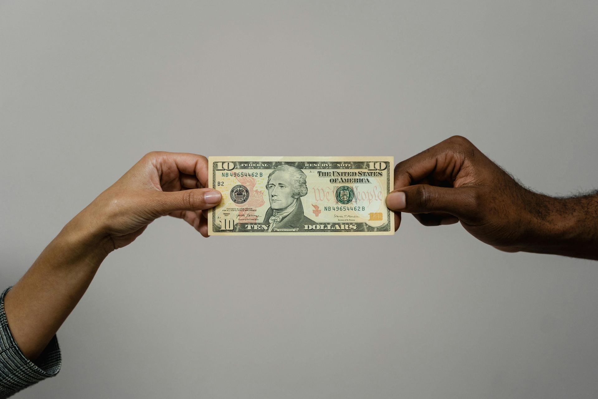 Two hands exchanging a U.S. dollar bill against a neutral background, symbolizing financial responsibility and the principle “You can’t manifest what you don’t manage.”