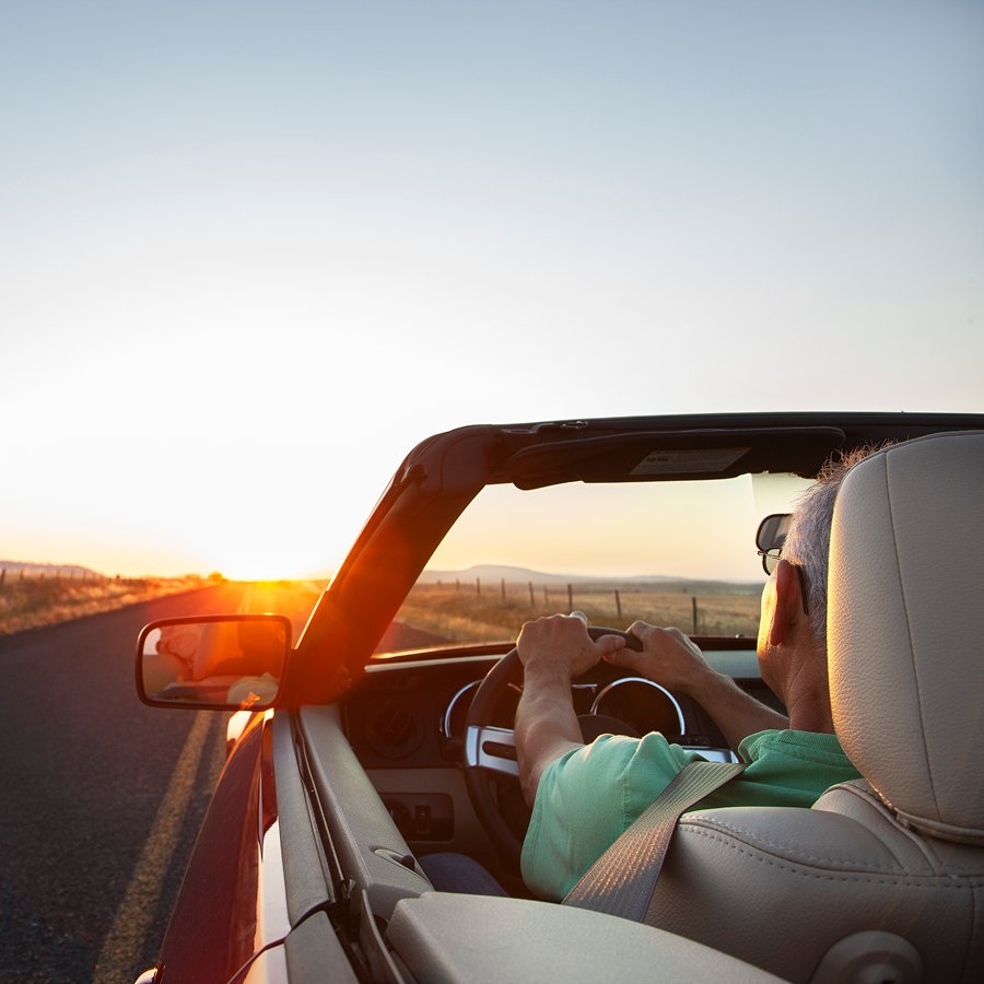 A man is driving a convertible car down a road at sunset