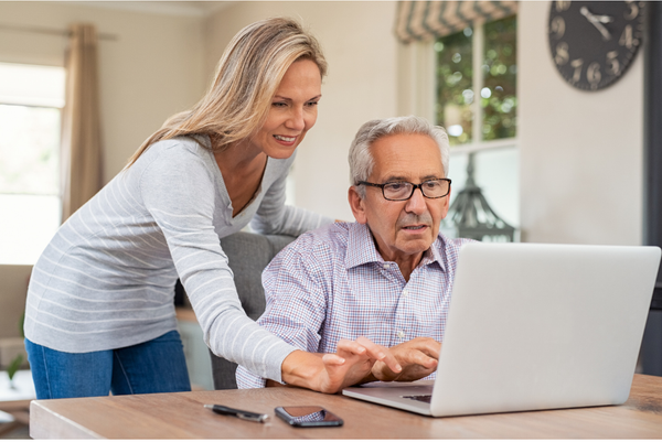 A picture of an older man on a laptop and a woman standing behind him helping him,