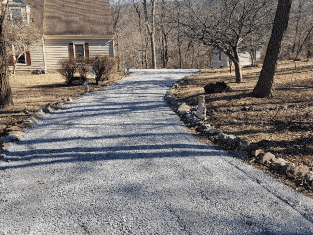 Gravel driveway leading up to a house, surrounded by trees and dry grass in a sunny setting.