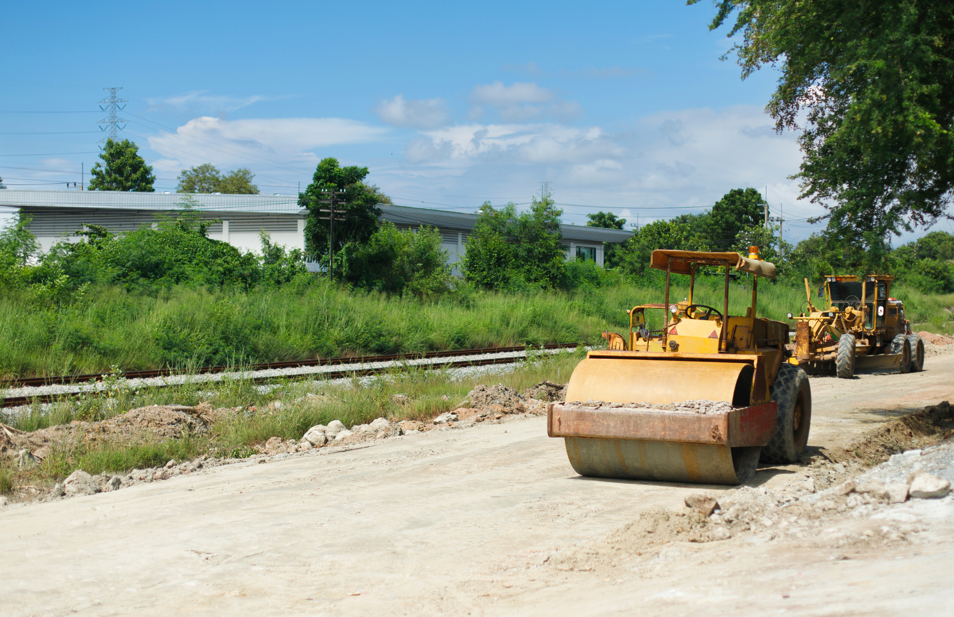 Road construction site with yellow roller and grader; green grass and buildings in background.