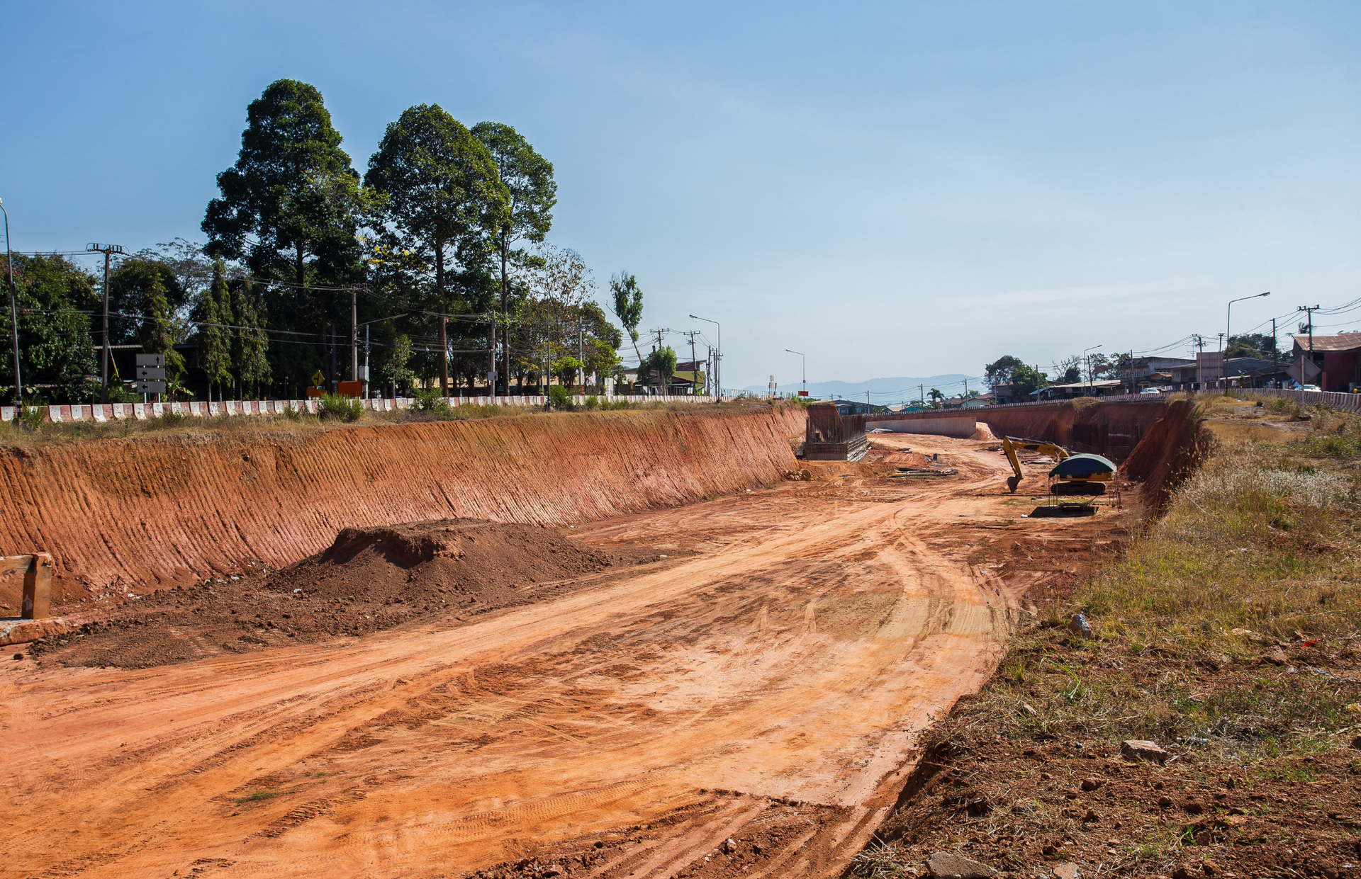 Two yellow excavators working on a large, brown construction site with a small body of water; houses and trees in background.