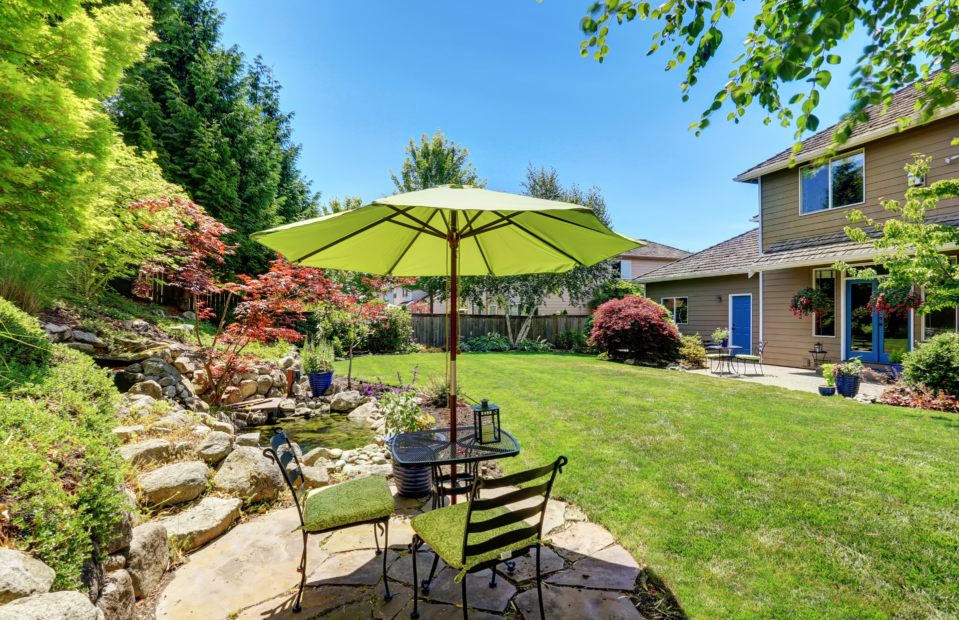 A sunny backyard with patio furniture under a lime green umbrella, near a small pond, and a two-story house.