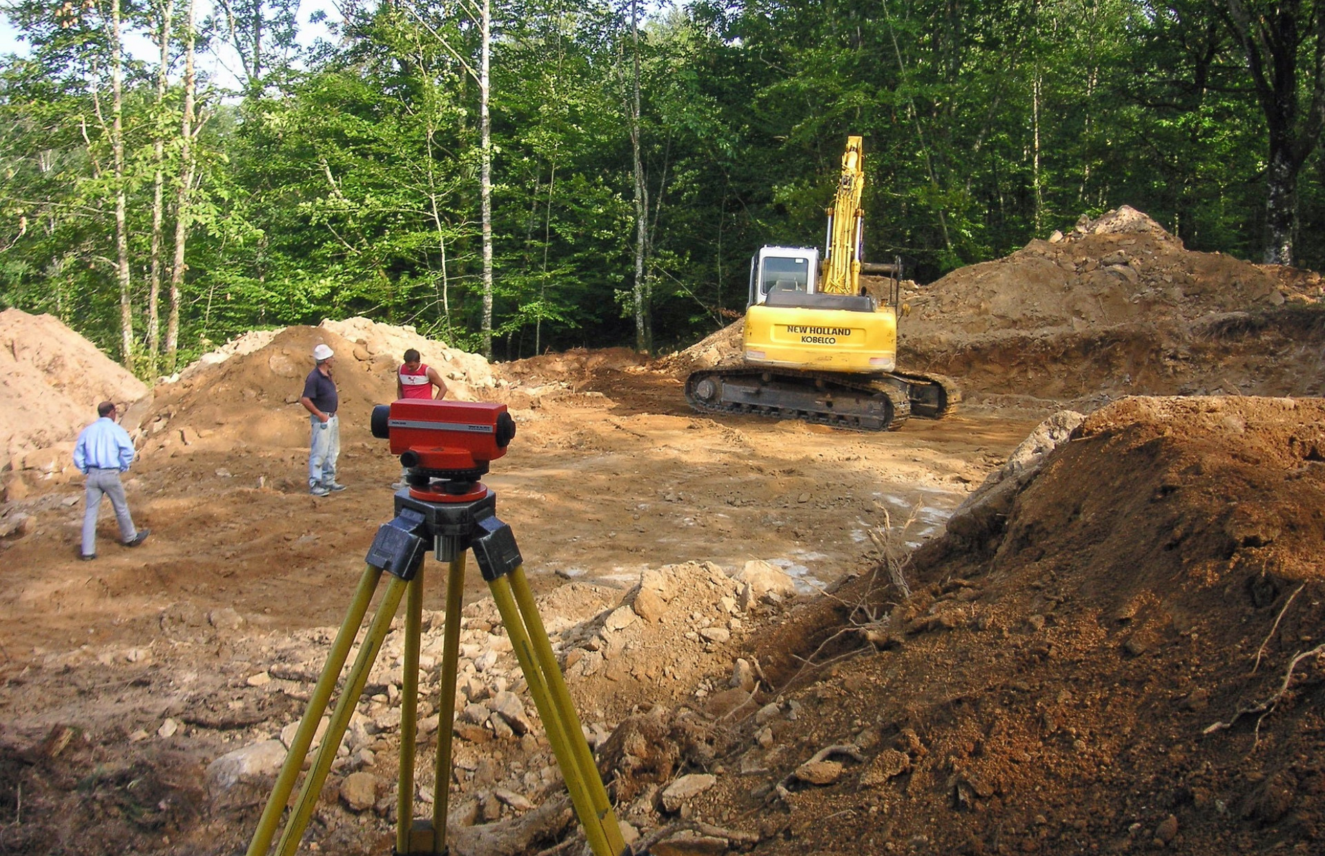 Construction site with surveyor's equipment, excavator, and workers on a wooded plot.