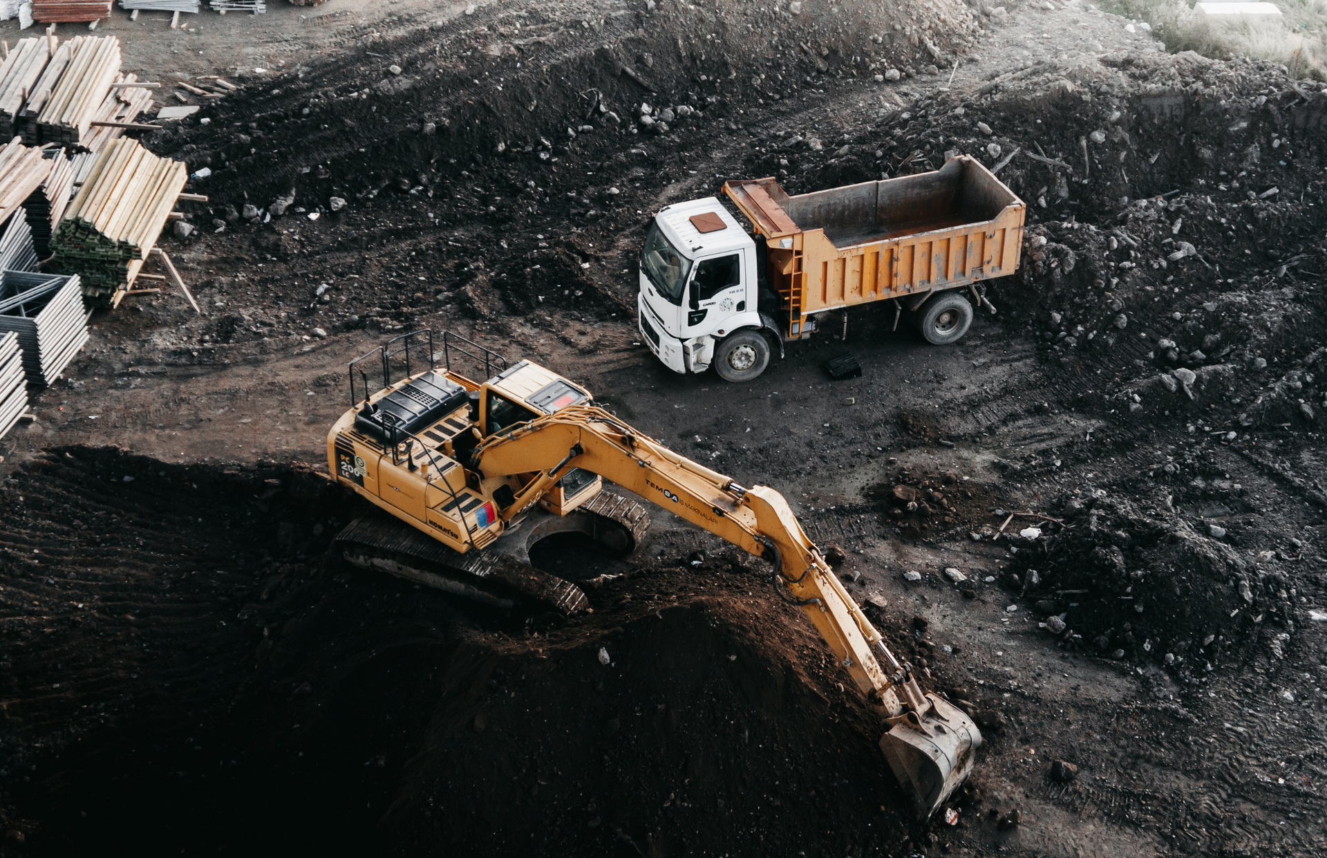 Three yellow dump trucks and an excavator in a quarry, hauling dirt and rocks, gray overcast sky.