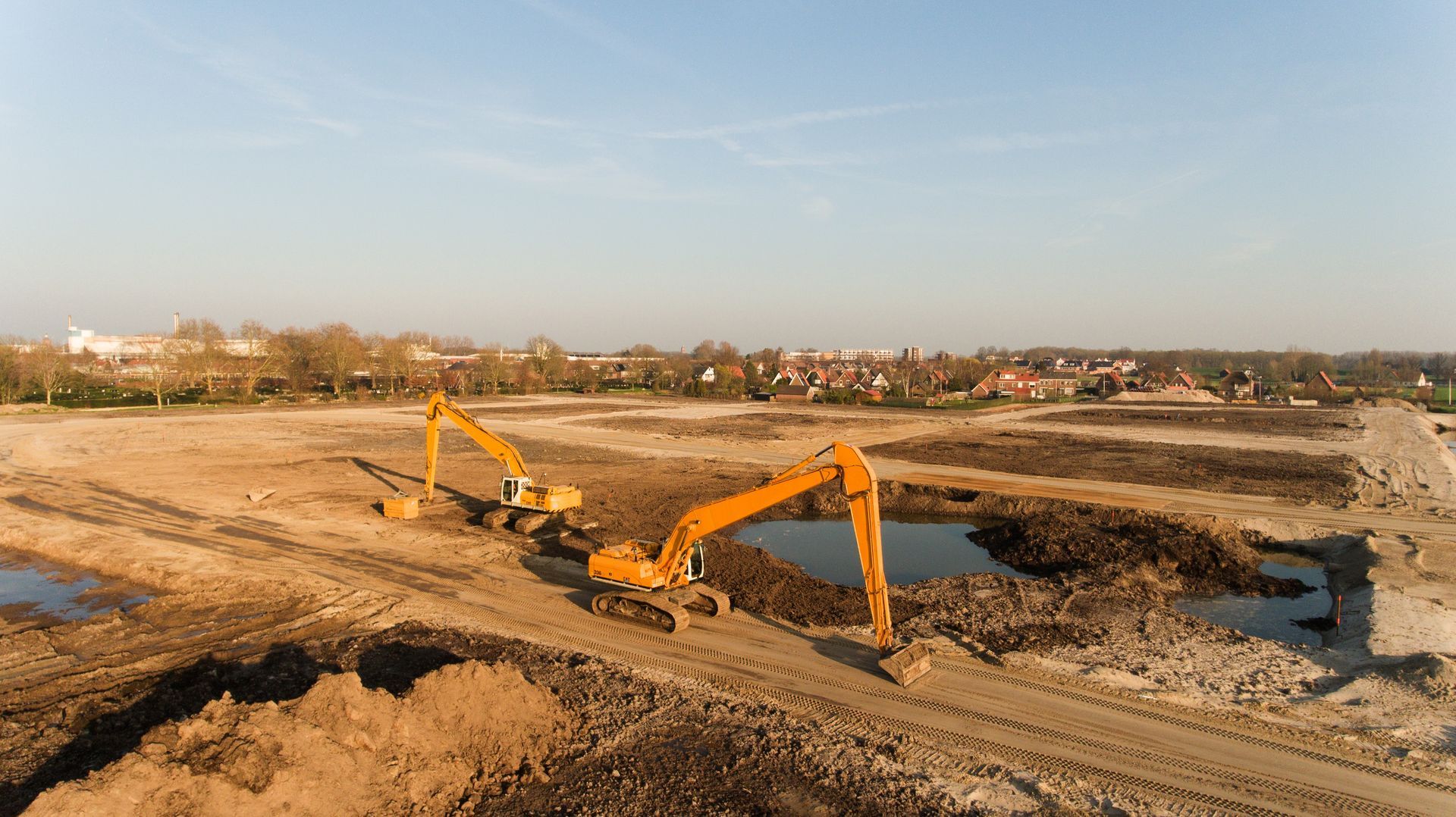 Two yellow excavators working on a dirt field, a residential area visible in the distance under a blue sky.