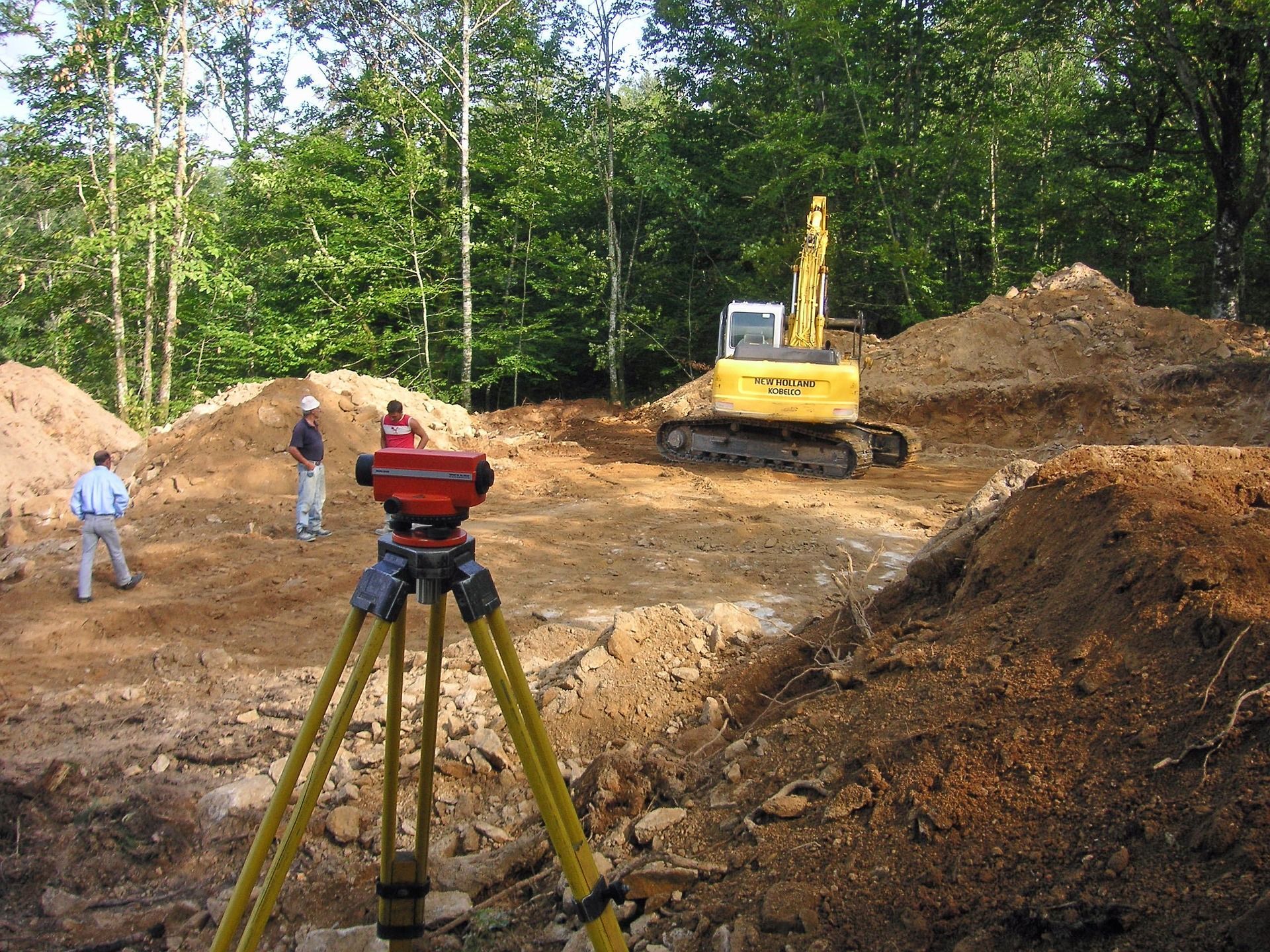Surveying equipment on tripod at a construction site, excavator, men in background, forest.