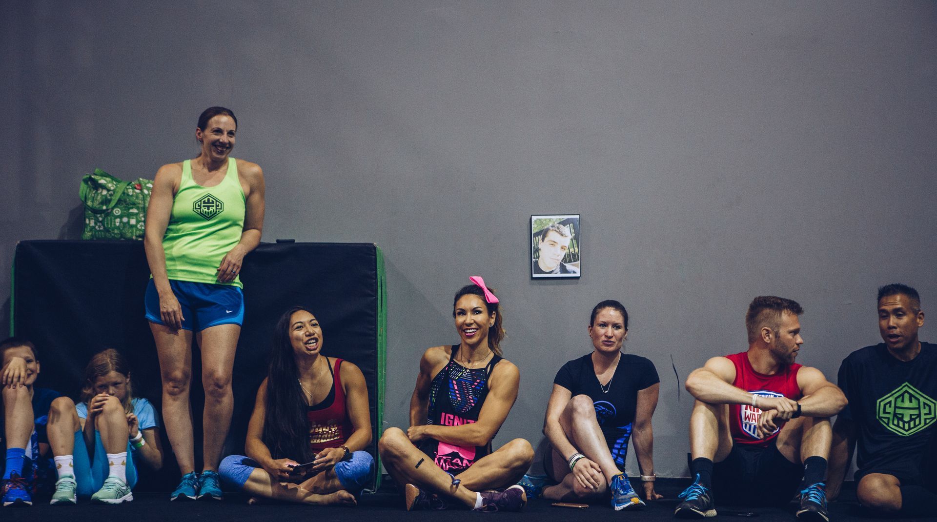 People sitting and standing against a gray wall in a gym. Some smile or laugh, wearing workout clothes.