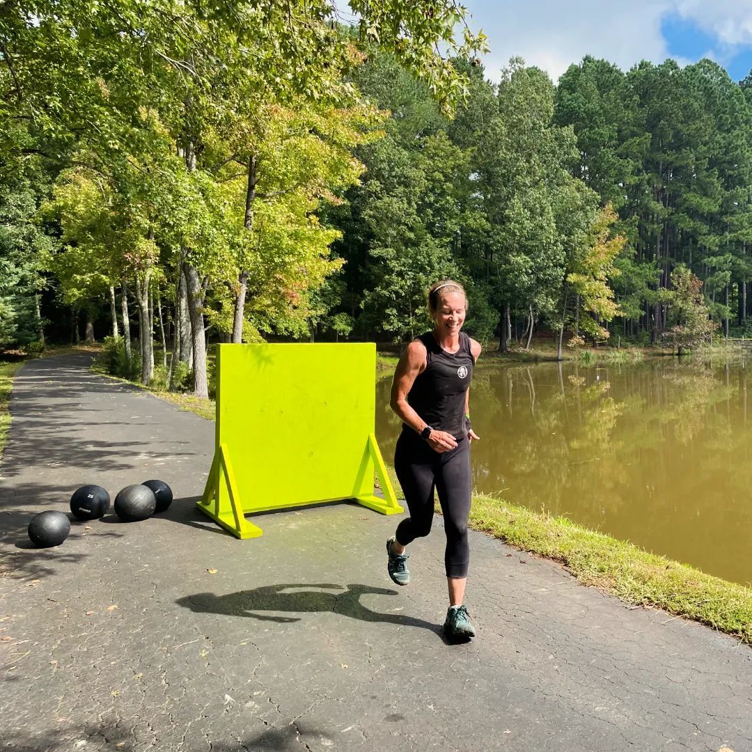 Woman running on a path beside a lake; green screen and medicine balls nearby.
