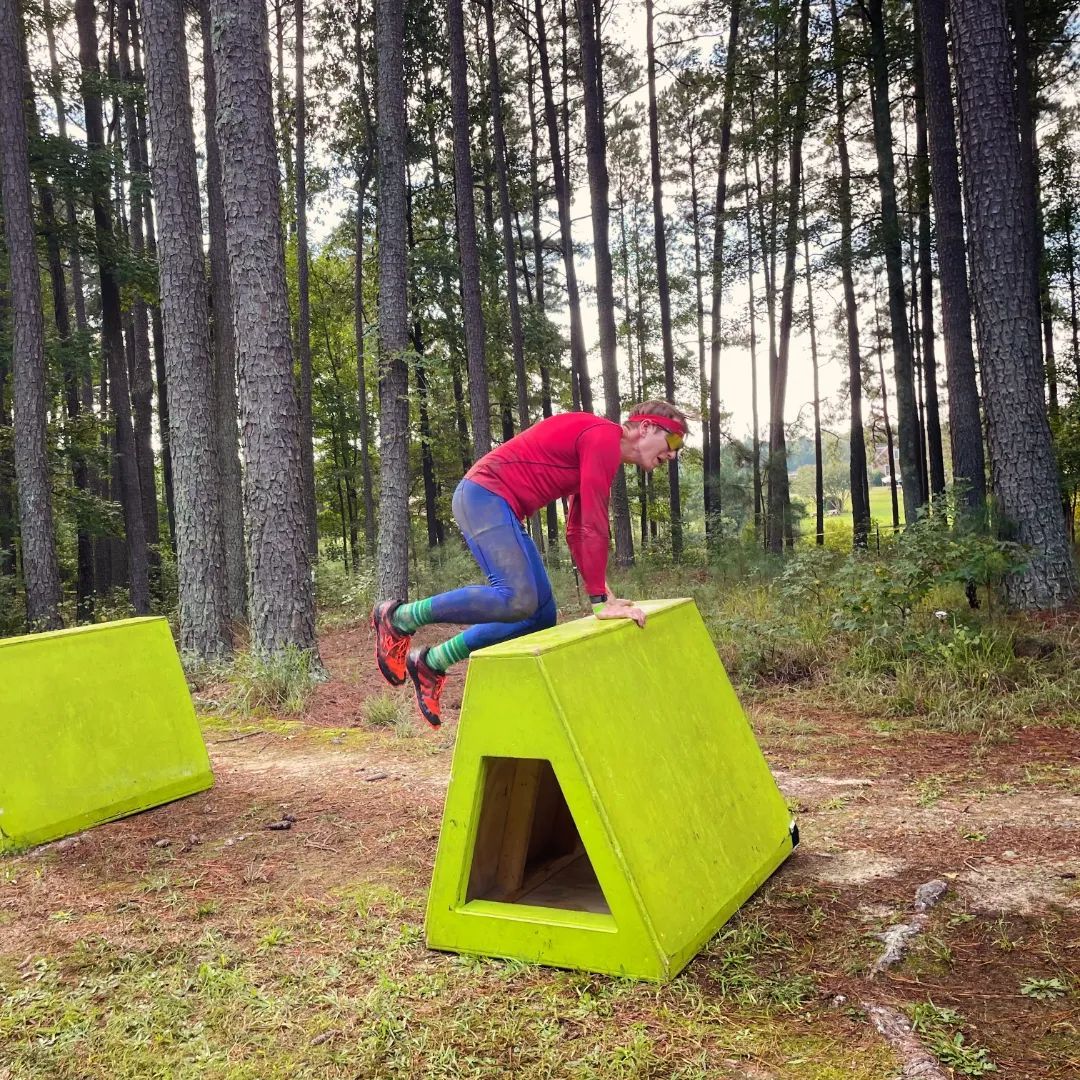 Person in red shirt and blue leggings jumps over a lime green obstacle in a forest setting.