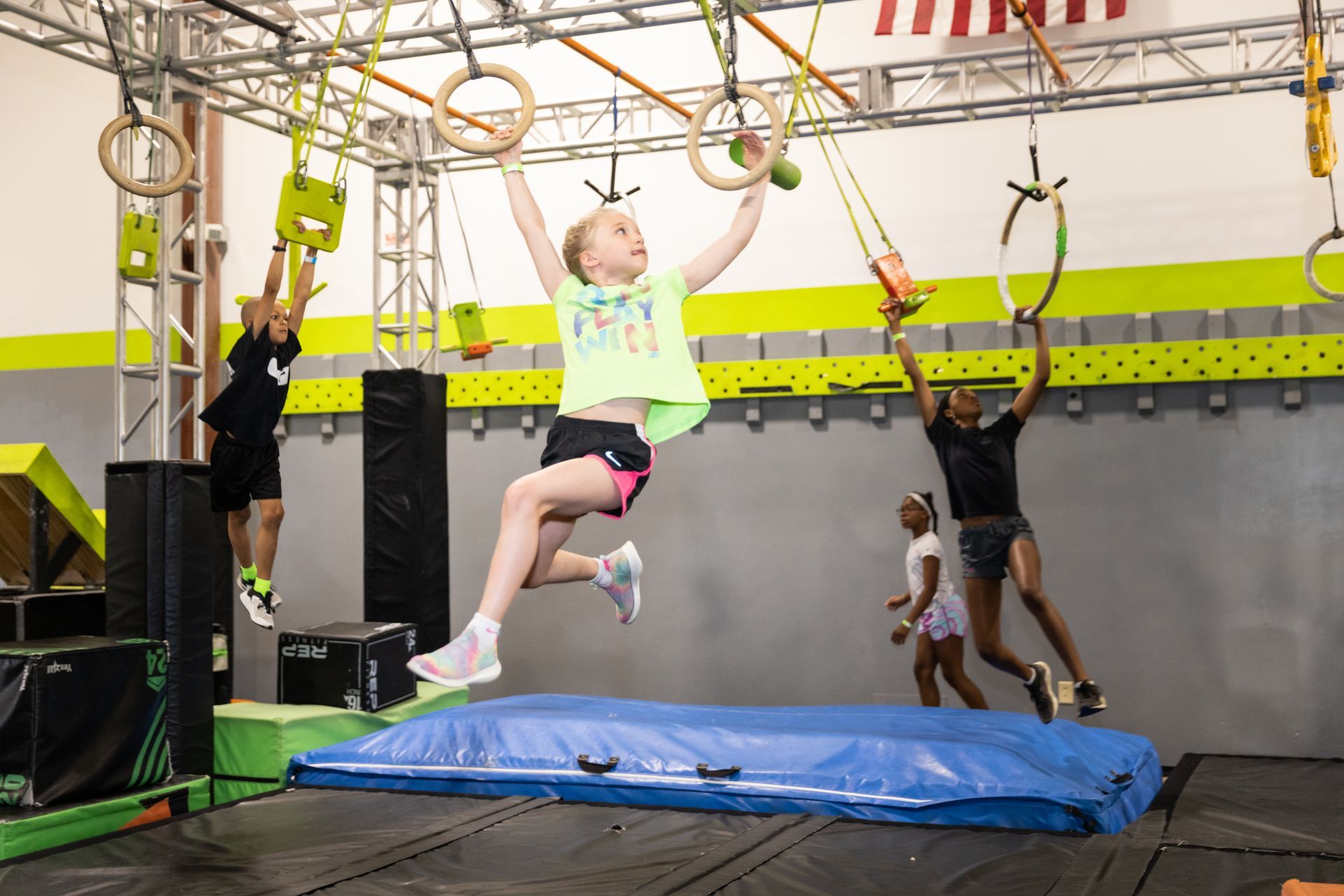 Children on an obstacle course, swinging on rings and bars. Indoor gym setting with mats and equipment.