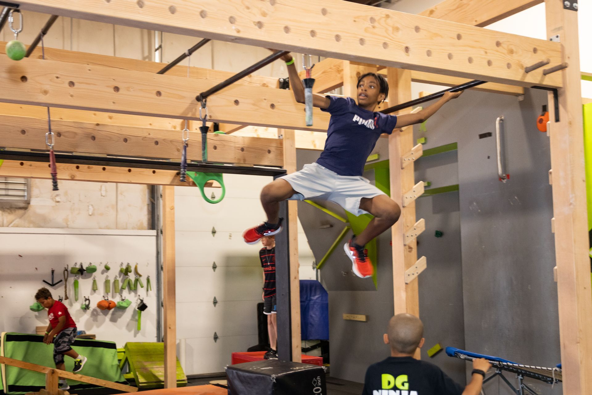 Person in mid-air on ninja obstacle course, legs spread, grasping overhead bars. Indoor gym setting.