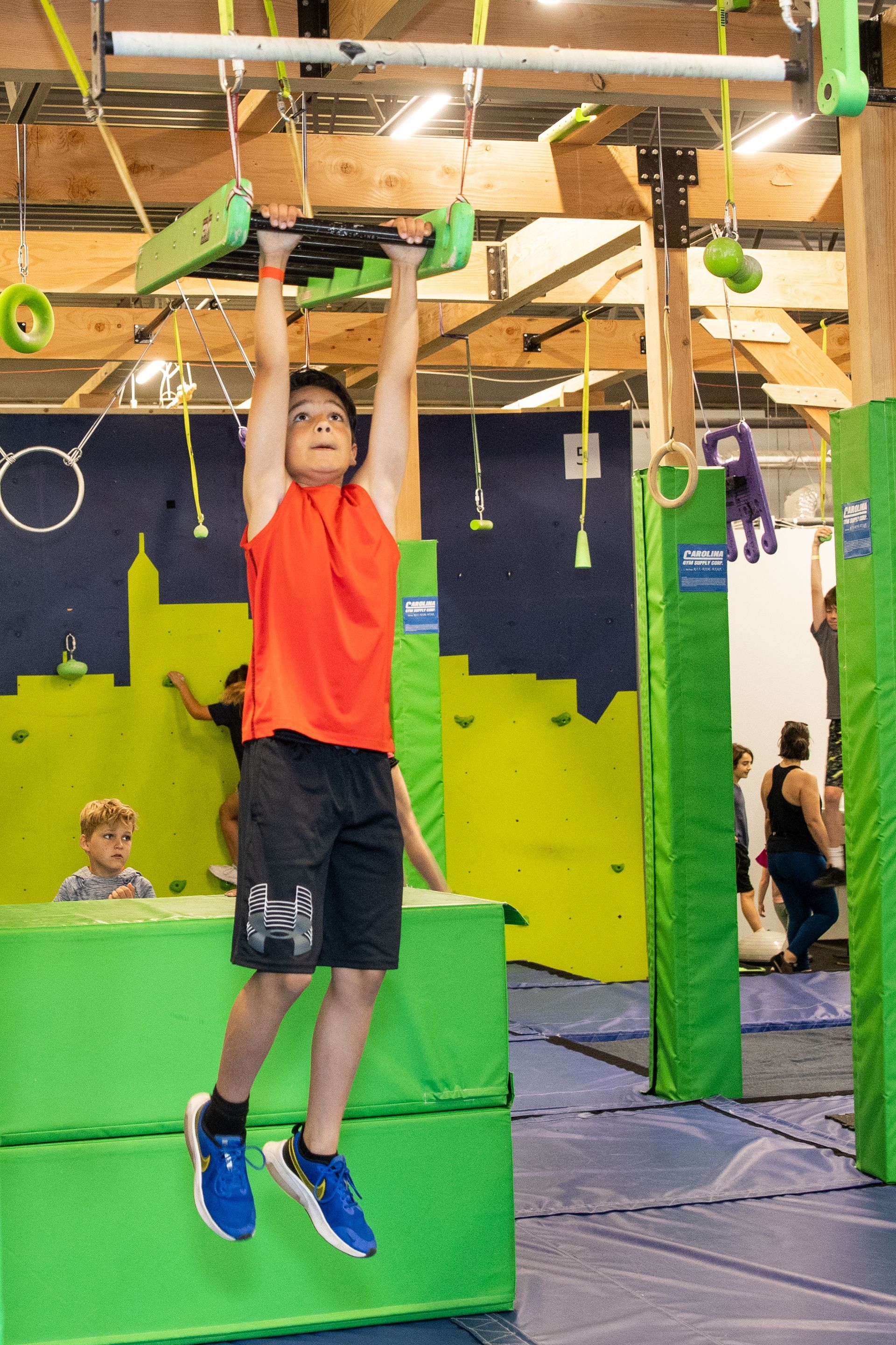 Boy in red shirt hanging from a bar at an obstacle course gym.