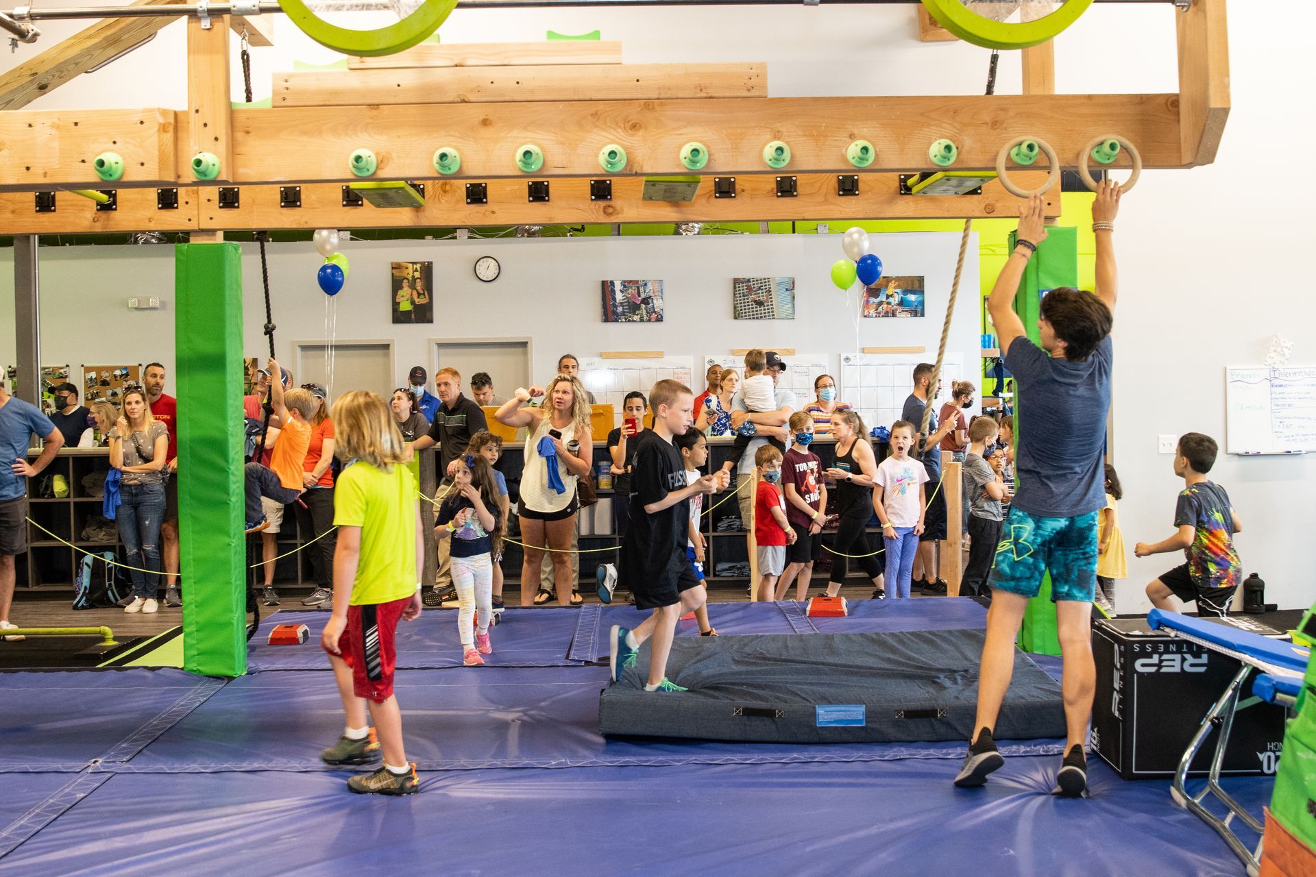 Children in a gym compete on an obstacle course, with a crowd of people watching.