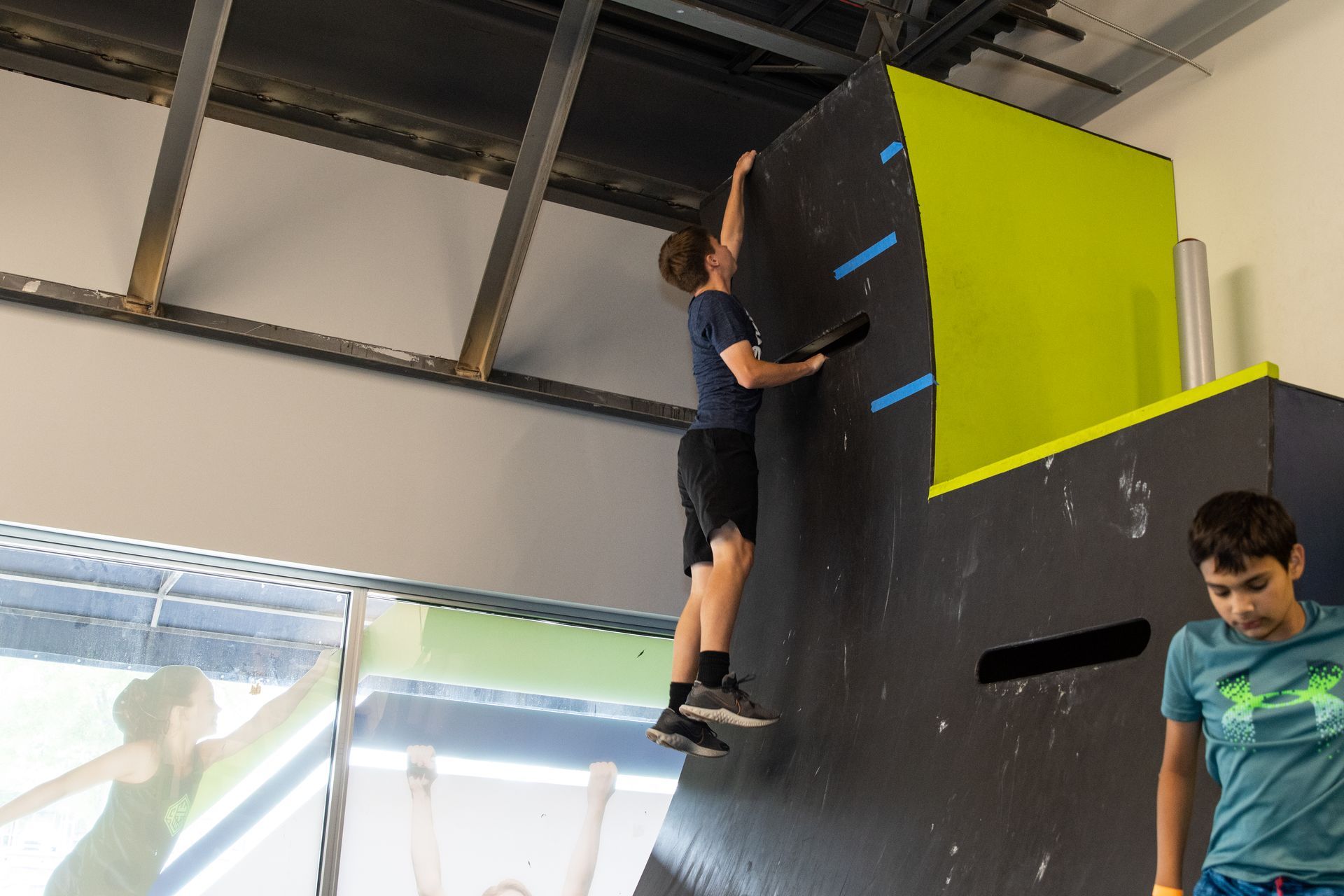 Boy reaching up a climbing wall in a gym; another boy watches.