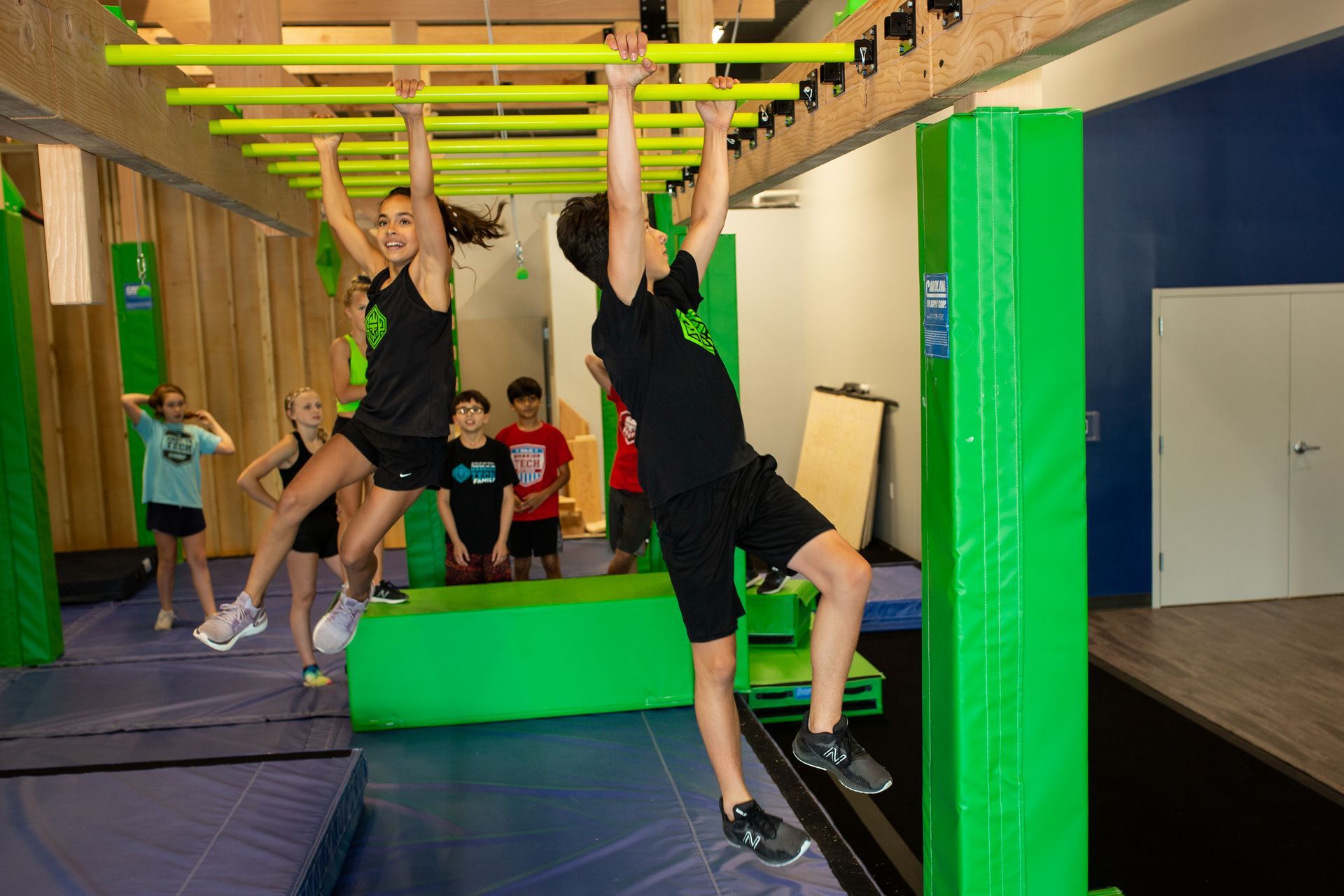 A boy and a girl are hanging on a monkey bars in a gym.