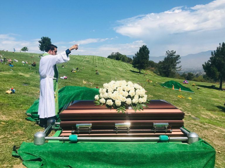 Catholic priest standing over casket with white flowers outside Chavez Funeral & Cremation Services, Inc. CA