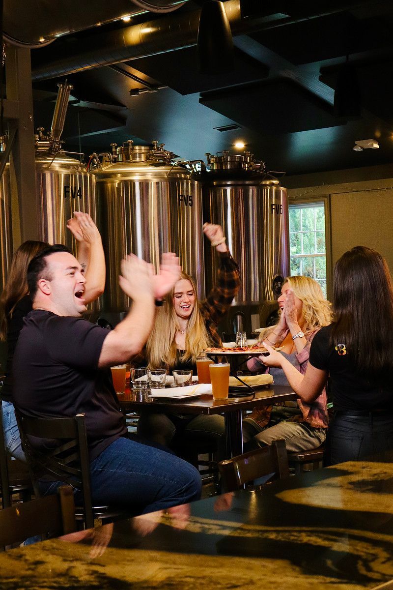People cheering at Brewbury, with large silver tanks in the background and food on the table.
