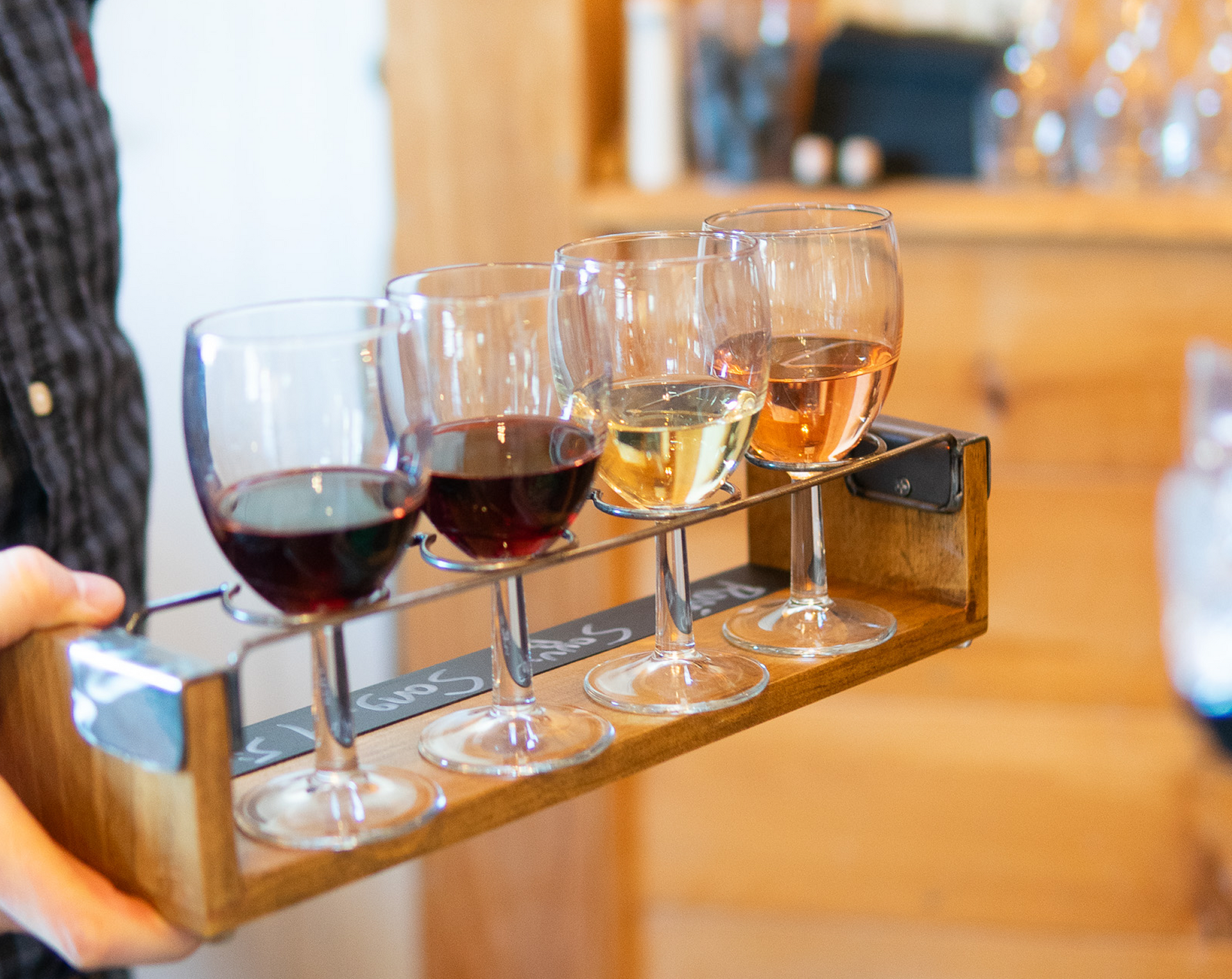 Person holding a wooden tray with four wine glasses containing red, white, and rose wine at Tranquillity Vineyard.