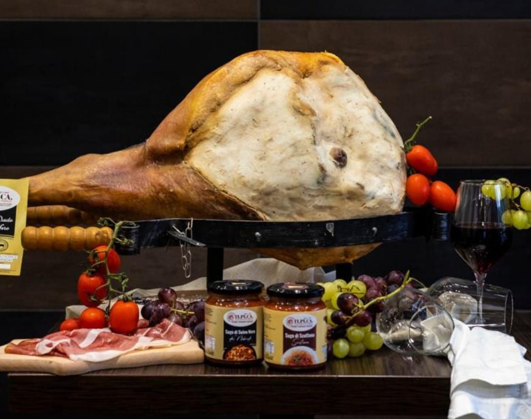 Un grande prosciutto è appoggiato su un tavolo, accanto a barattoli di salsa e uva.