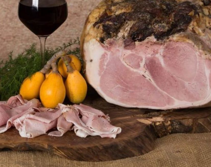 Un grosso pezzo di prosciutto è appoggiato su un tagliere di legno accanto a un bicchiere di vino.
