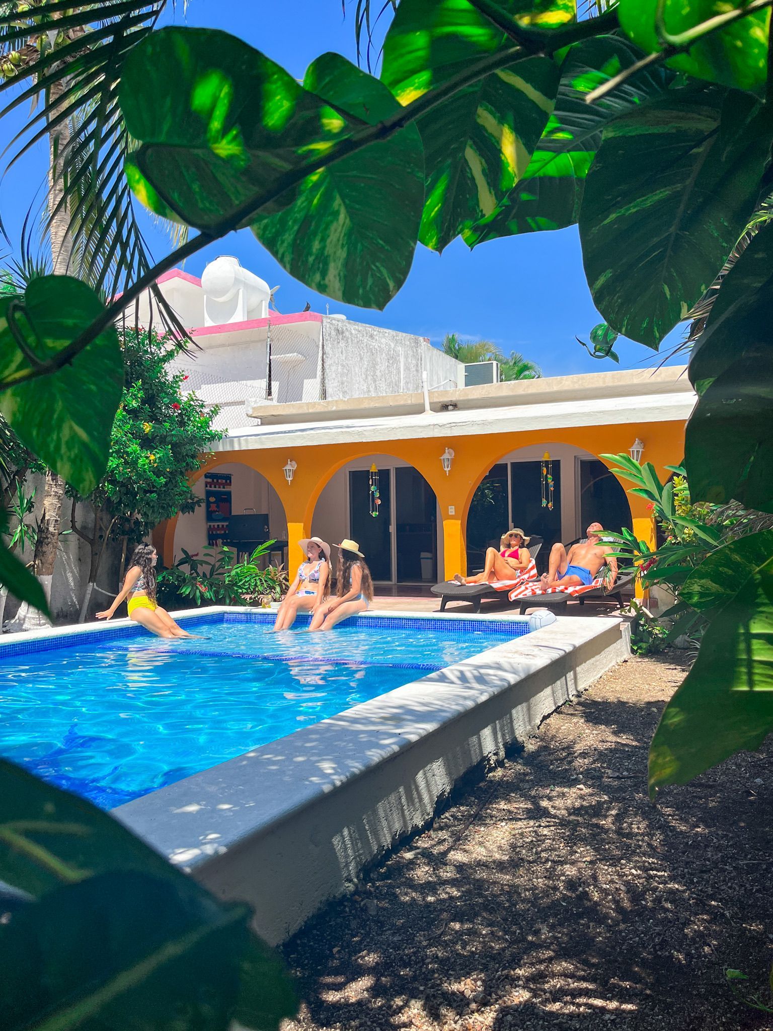 People relaxing by a pool in a tropical setting. Yellow building, blue water, green foliage, sunny day.