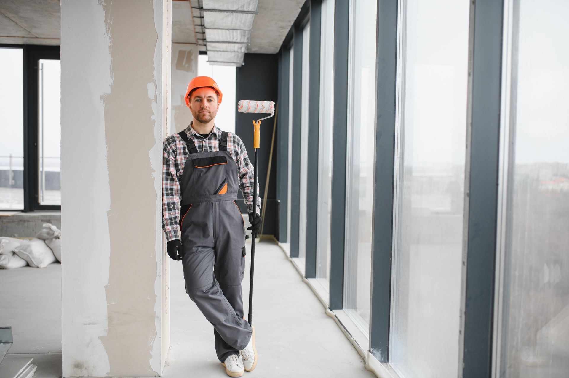 A male worker holding a paint roller near a window in a commercial building under construction.