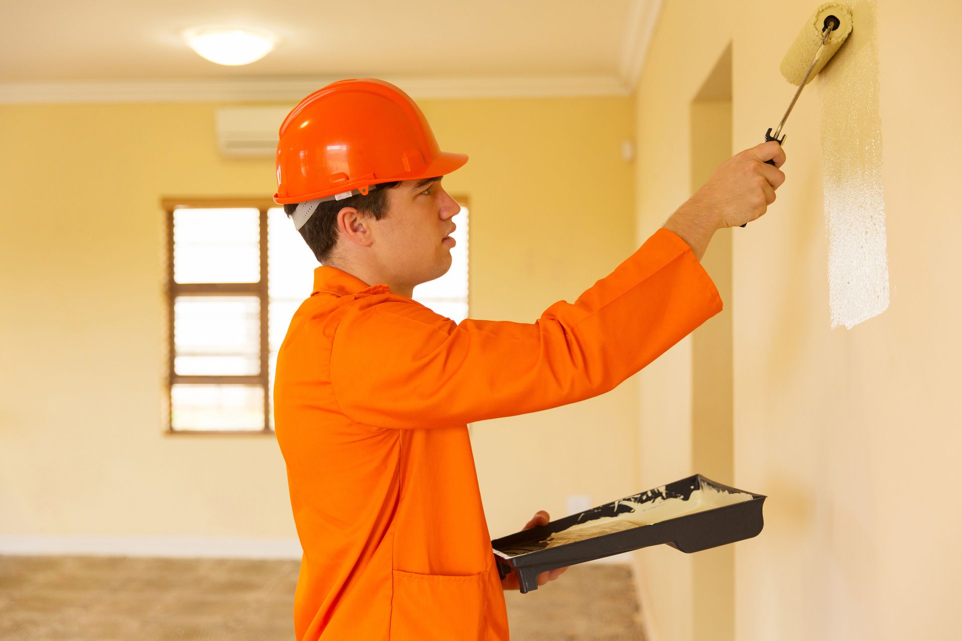 Worker in orange uniform painting beige wall with roller during interior renovation project.
