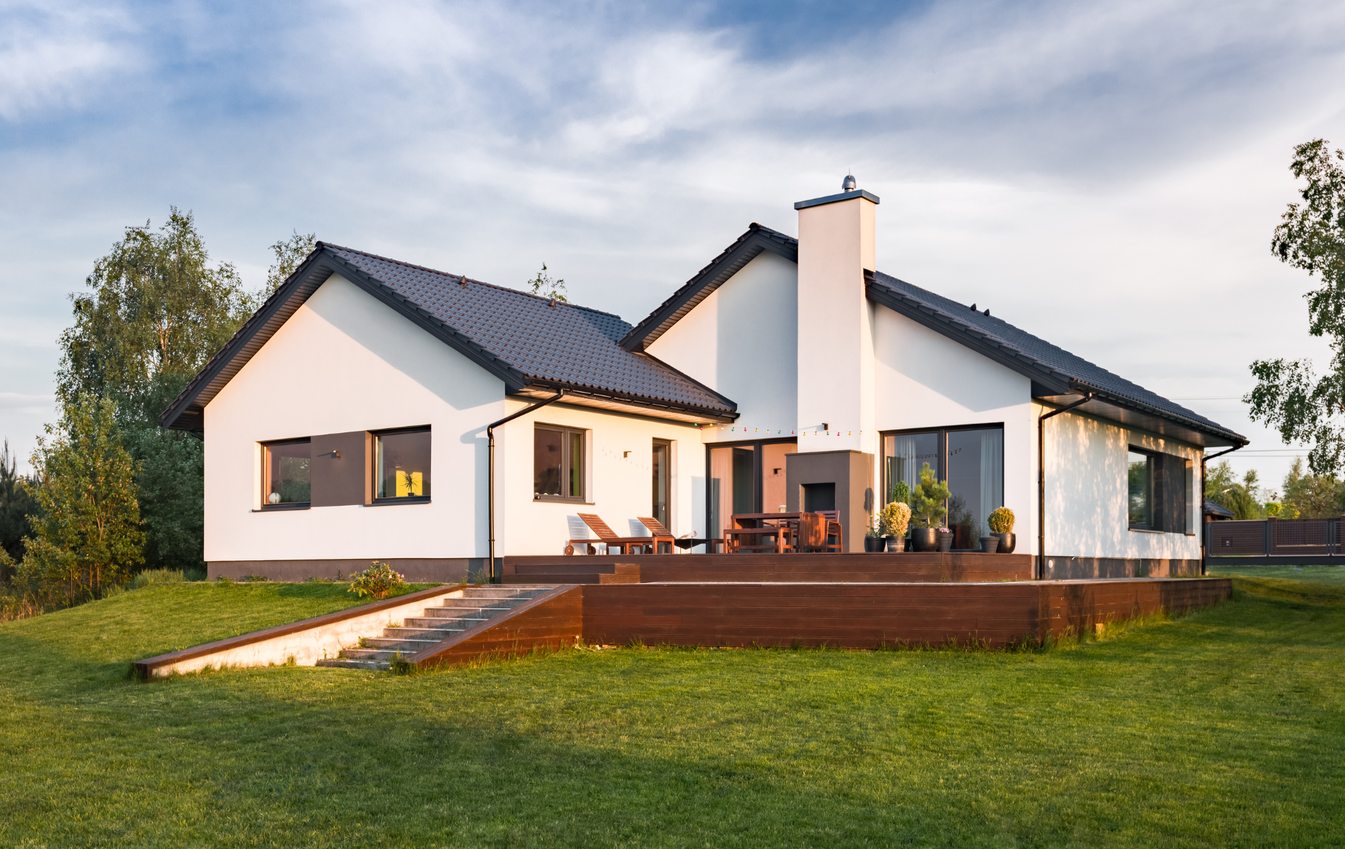 Modern white house with dark roof, wooden deck, chimney, and green lawn under a blue sky.