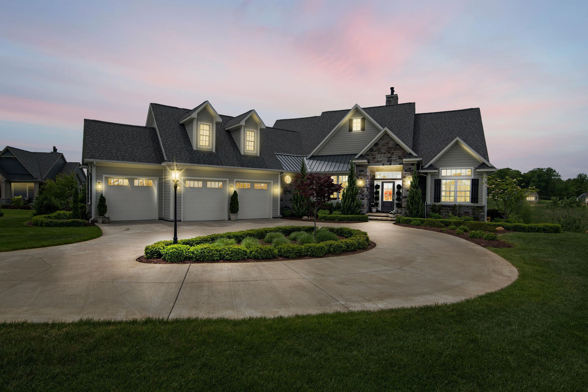 Large suburban house at dusk with lit windows, triple garage, and a circular driveway with landscaped island