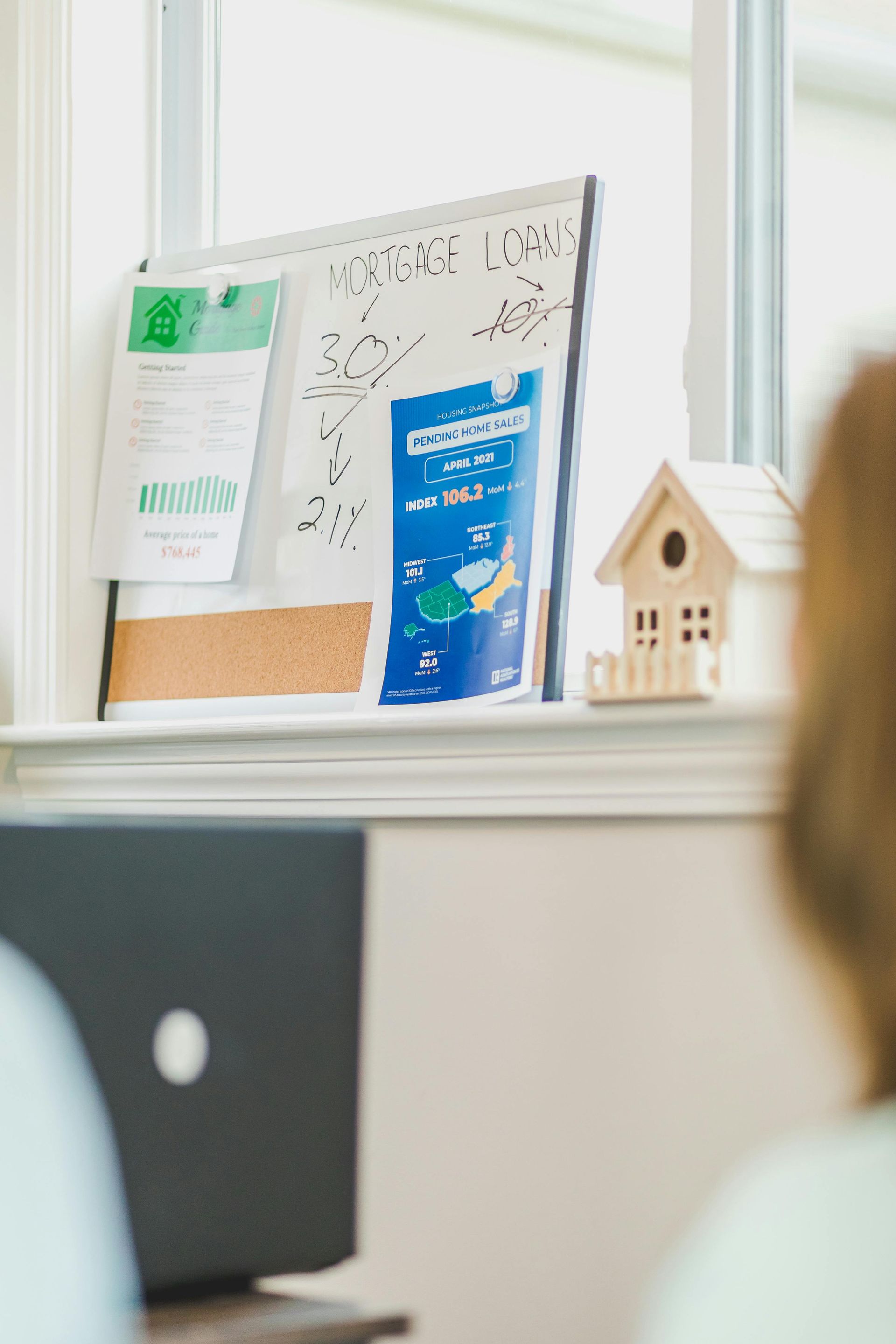 A whiteboard with notes and a blue flyer sits on a windowsill next to a small model house, with a laptop in the foreground.