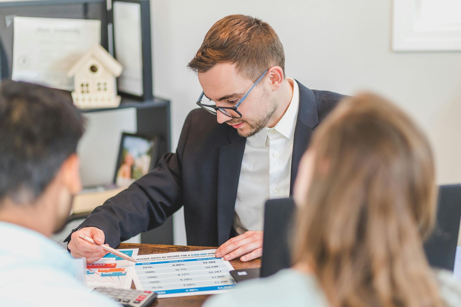 Man in suit reviews documents with couple at a desk, house model in the background.